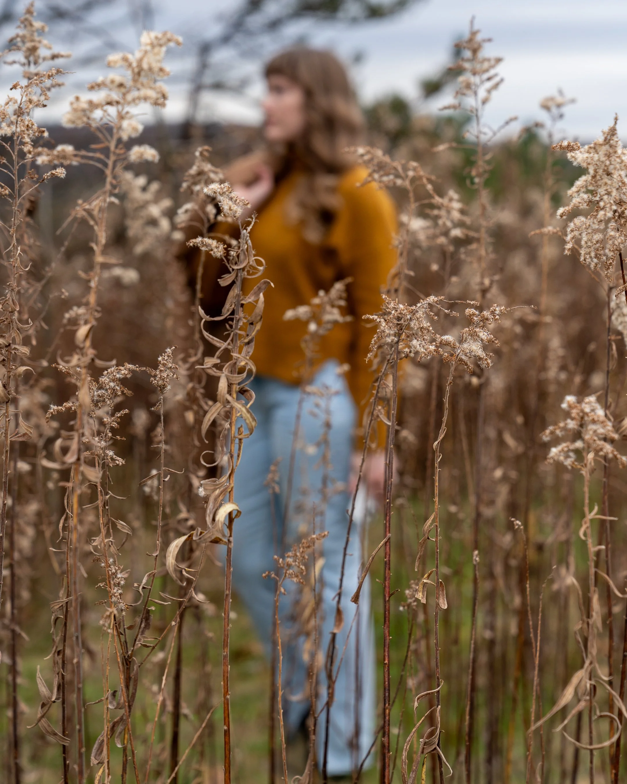 A woman with brown hair wearing a mustard yellow jacket and light blue jeans standing in a field of dried plants, with her face slightly out of focus in the background.