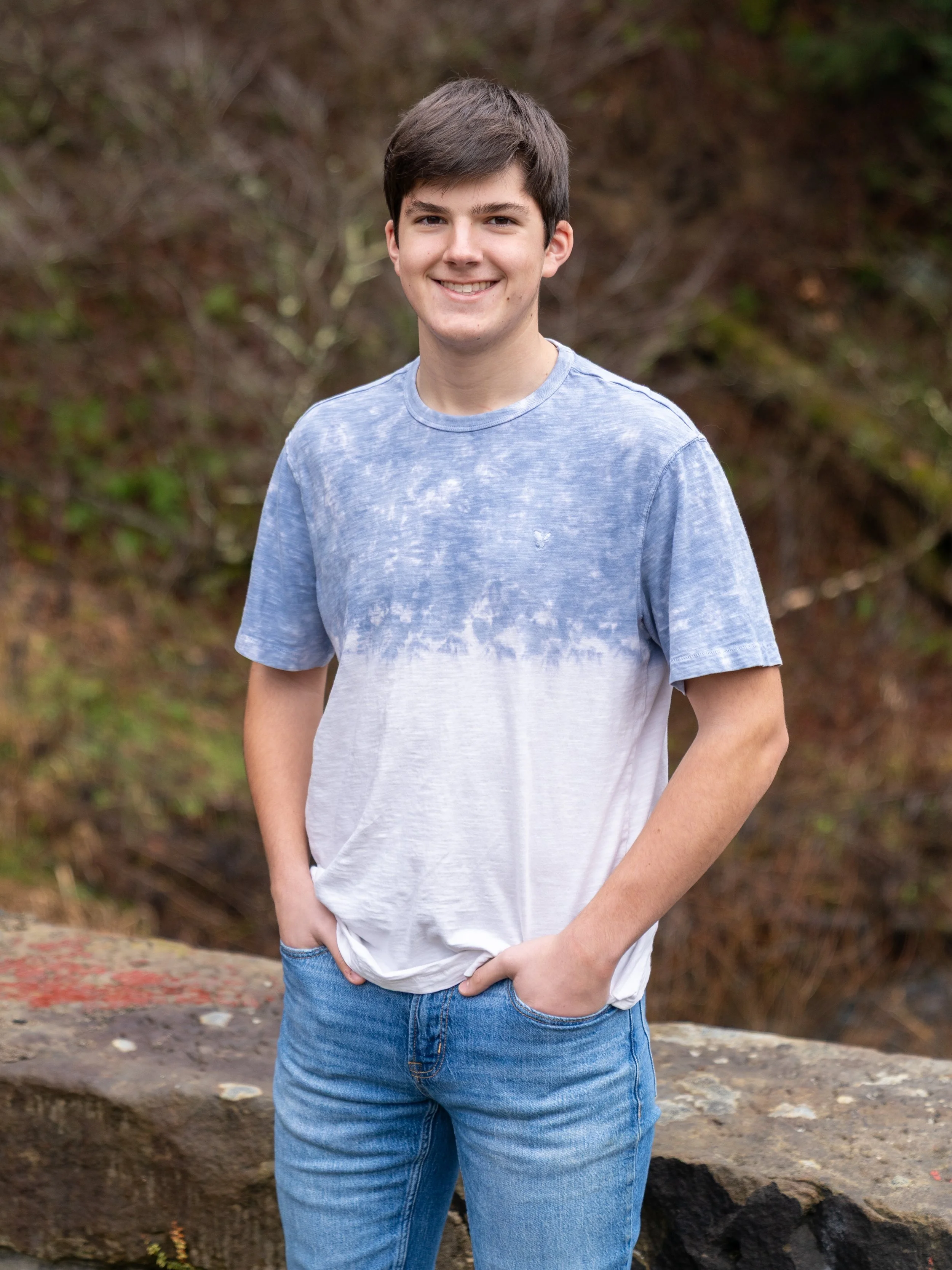 A young man in blue jeans and a blue and white tie-dye T-shirt standing outdoors in front of a blurred natural background, smiling and leaning slightly to one side.