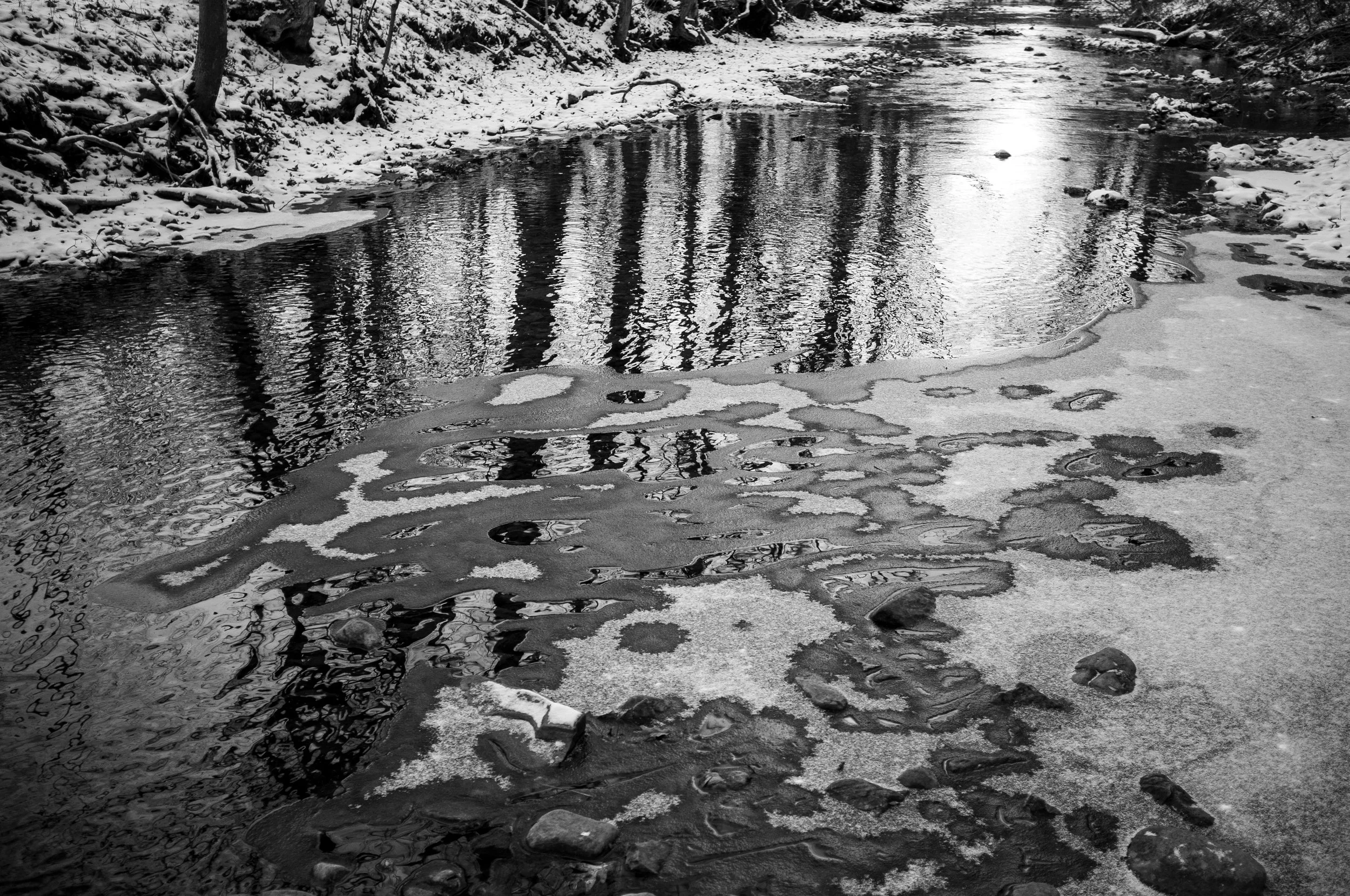 Frozen river with icy patches and snow-covered banks, leafless trees reflected on the water, in a winter forest scene.