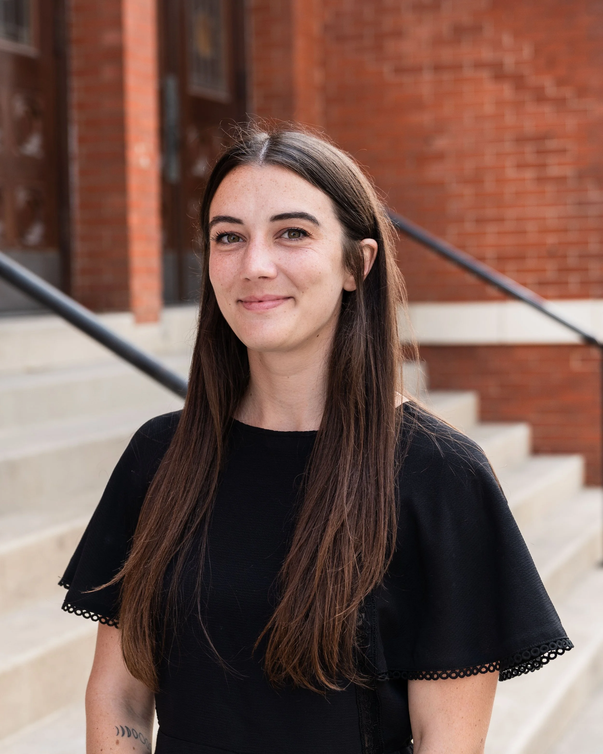 Close-up photo of a woman with long brown hair, wearing a black top with lace trim on the sleeves, standing outdoors in front of a brick building with stairs and railing.