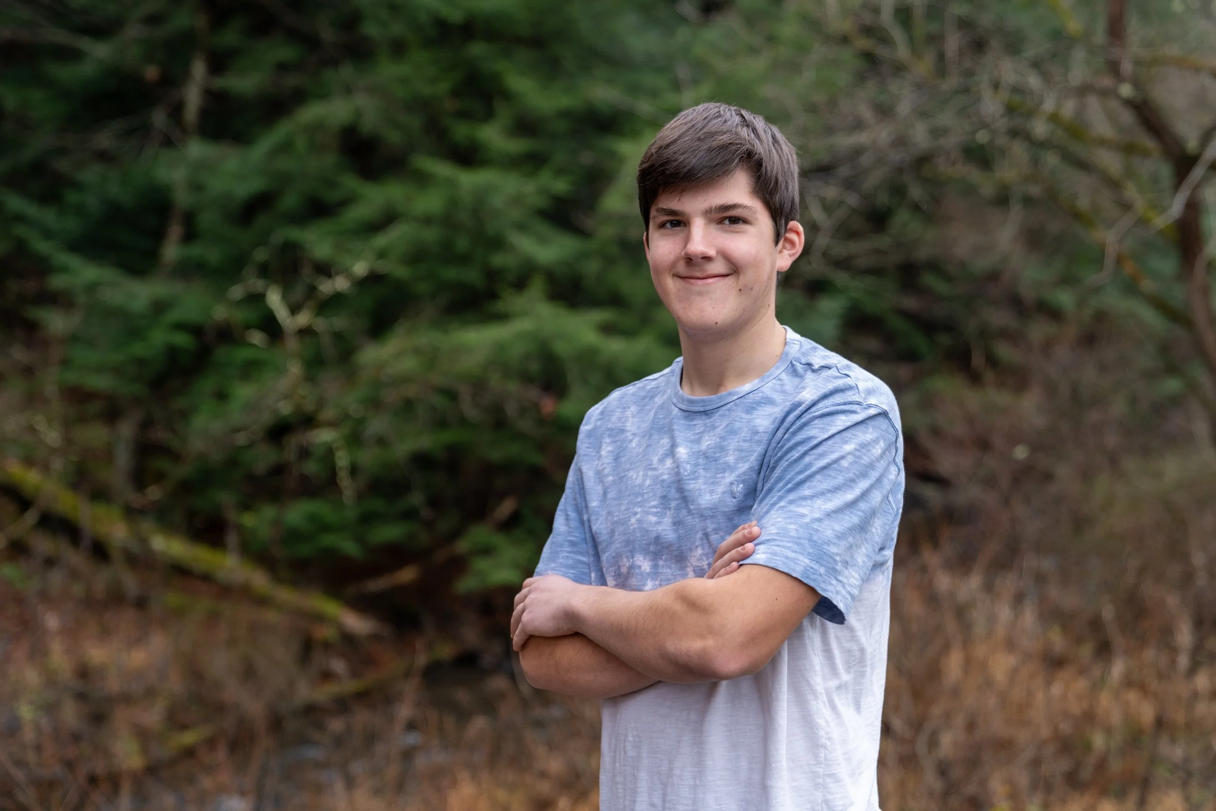A young man with dark hair smiling and crossing his arms outdoors with trees and foliage in the background.