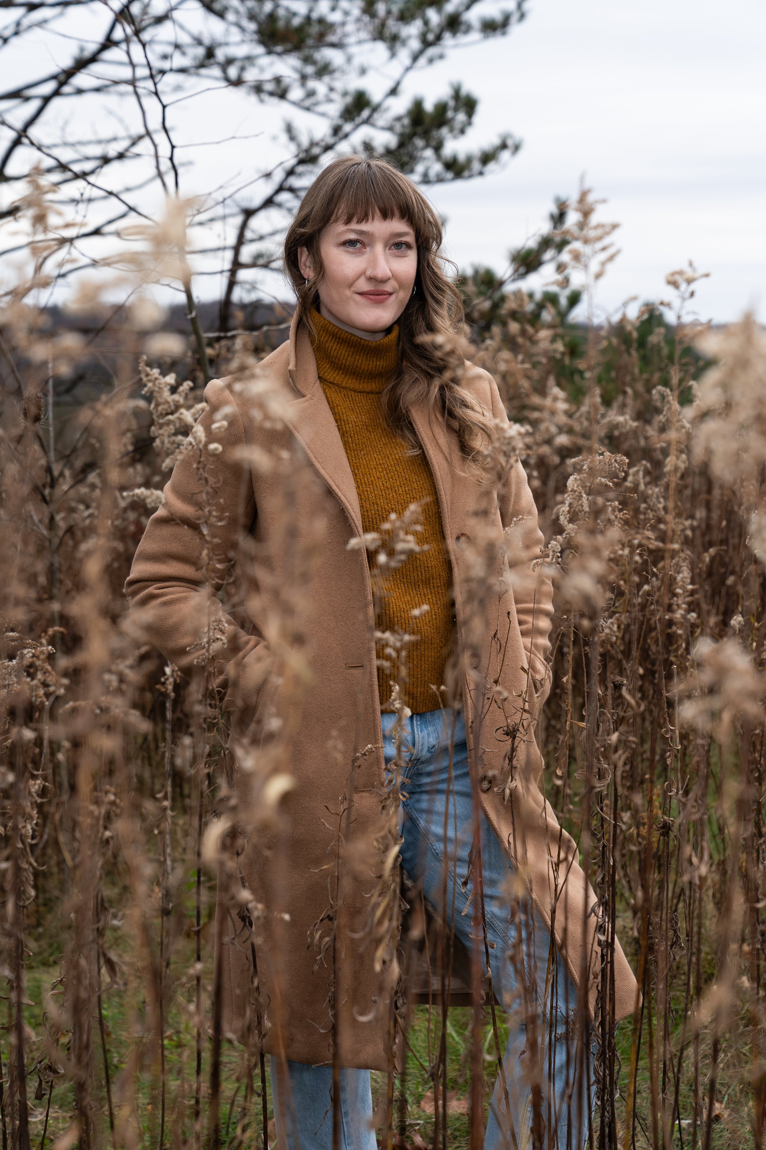 A woman with brown hair, wearing a tan coat and a mustard turtleneck sweater, standing in a field of dried plants with trees and an overcast sky in the background.