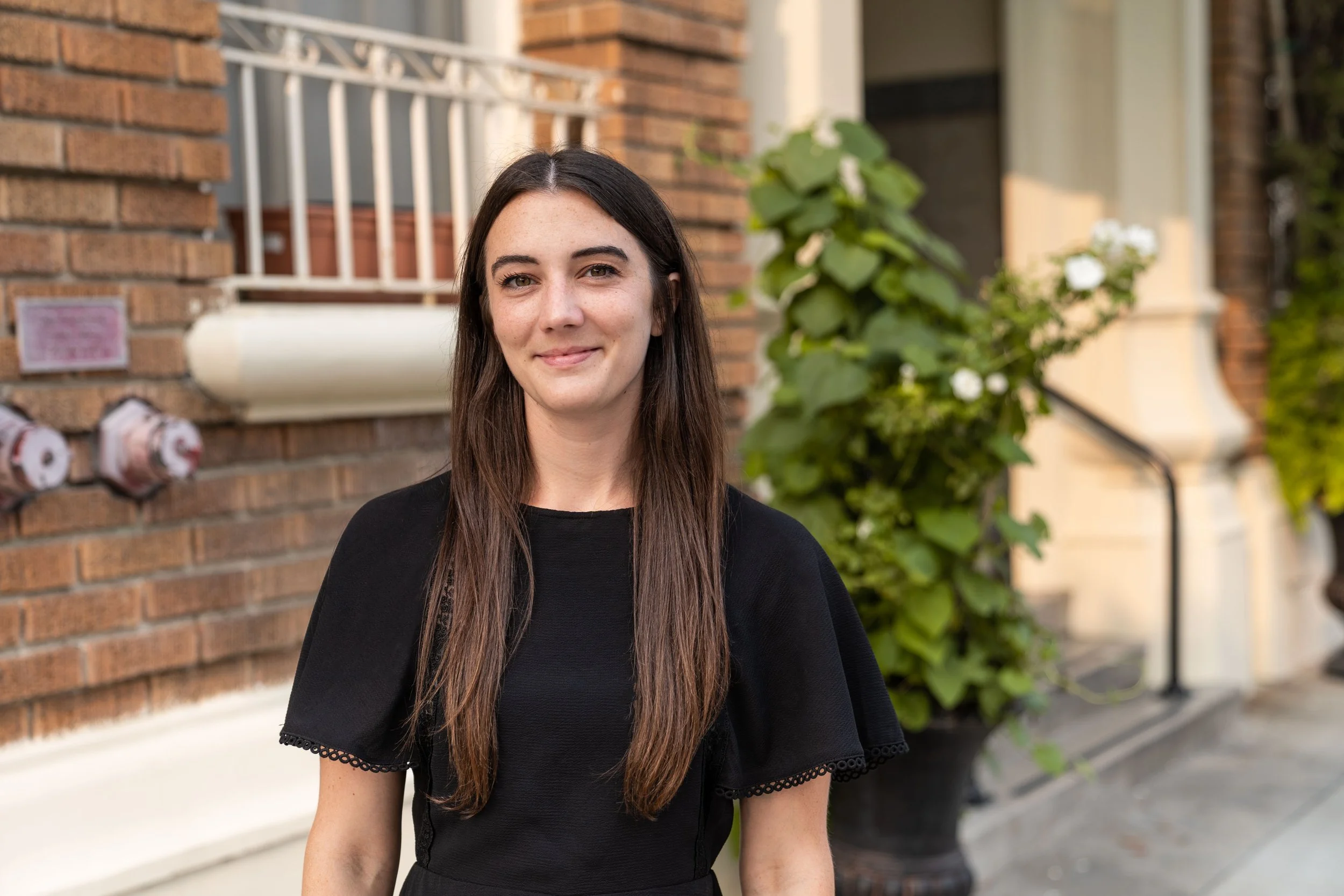 A woman with long brown hair and fair skin smiling outdoors in front of a brick building and greenery, wearing a black dress.