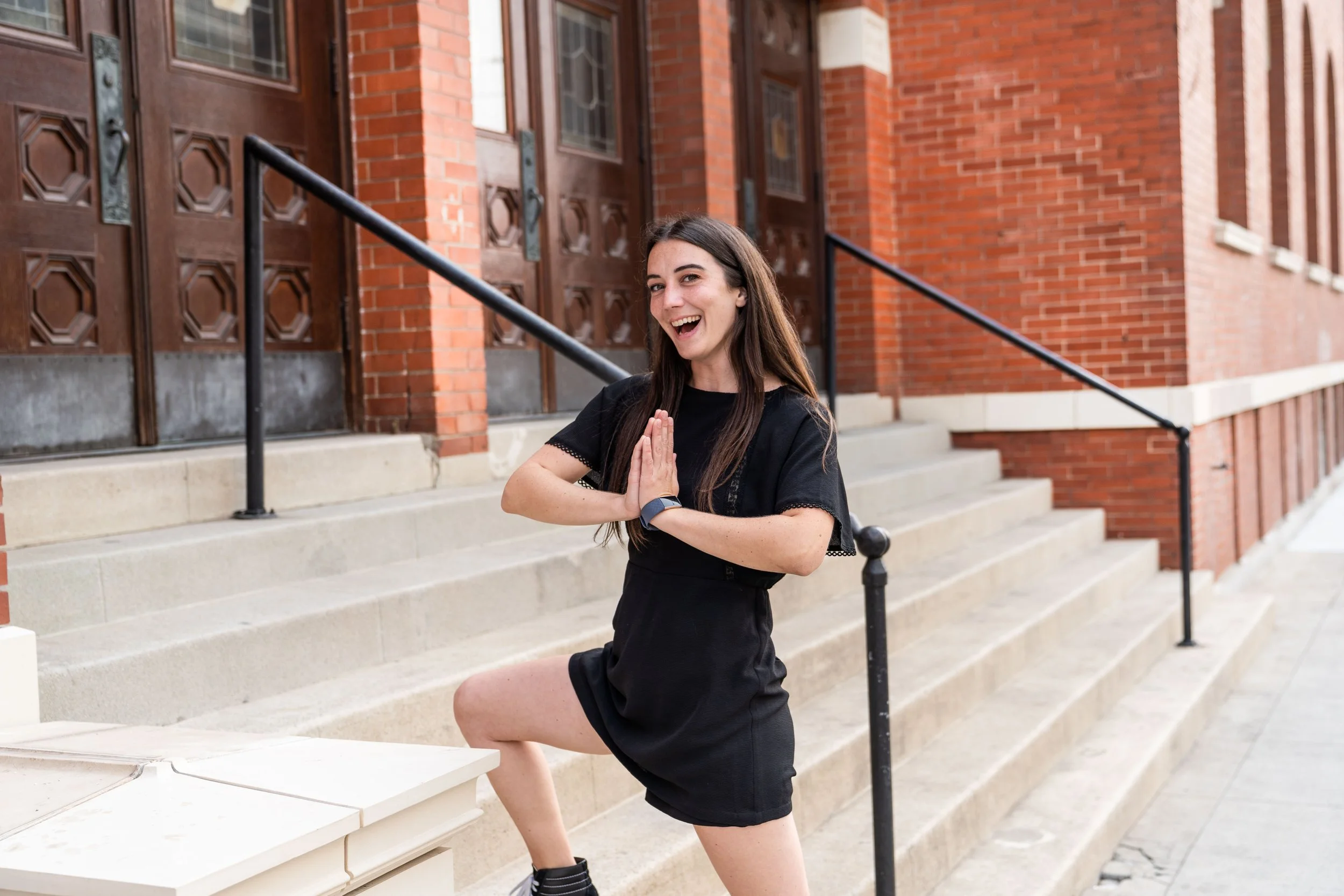 A young woman practicing yoga outdoors on stairs in front of a brick building, with one leg raised, hands in prayer position, and smiling.