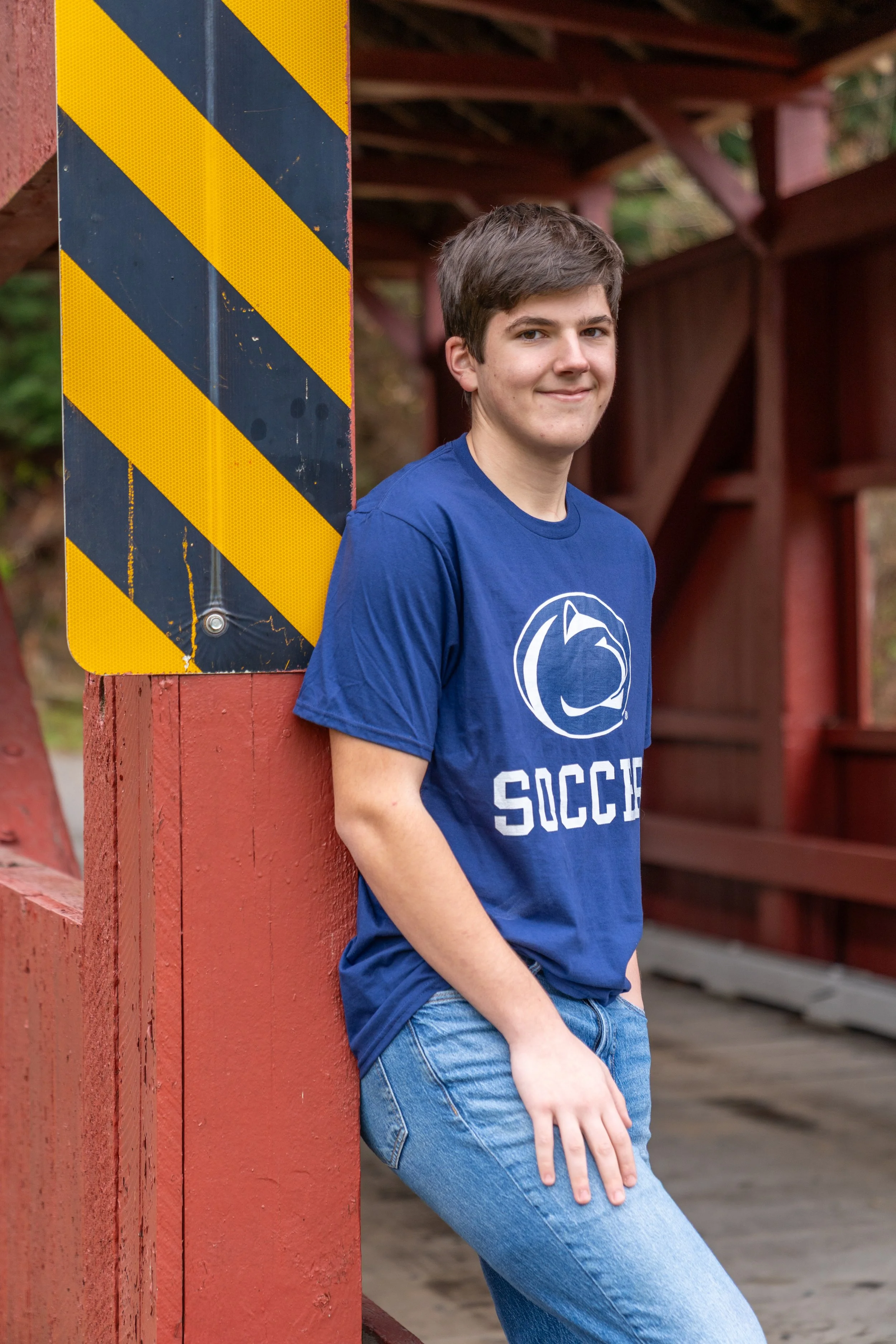 A young man with short brown hair wearing a blue t-shirt and jeans, standing outdoors by a red wooden structure with a yellow and black striped safety sign attached.
