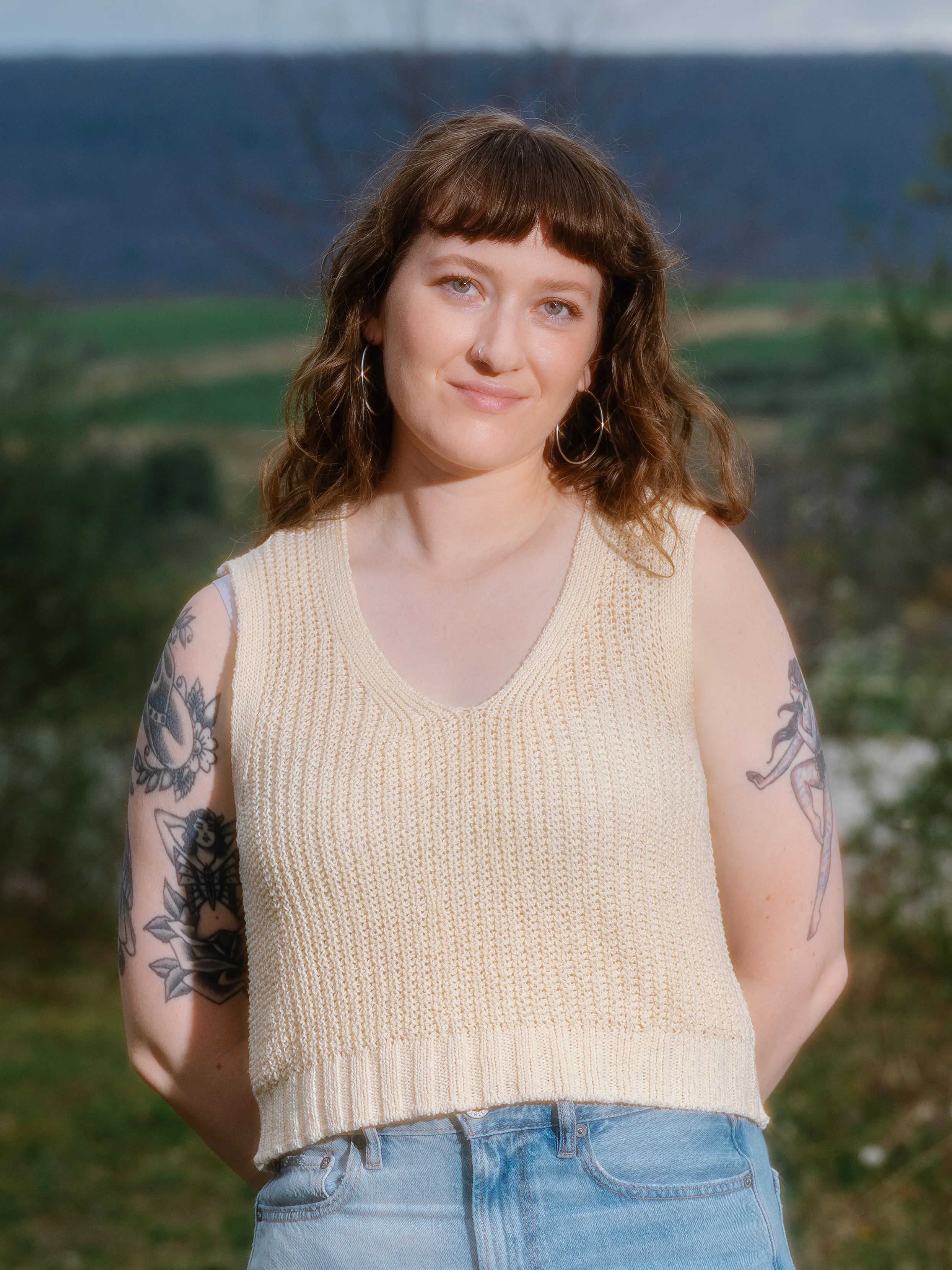 A woman with brown hair and blue eyes, wearing a sleeveless beige knit top, stands outdoors with a landscape of fields and mountains in the background.