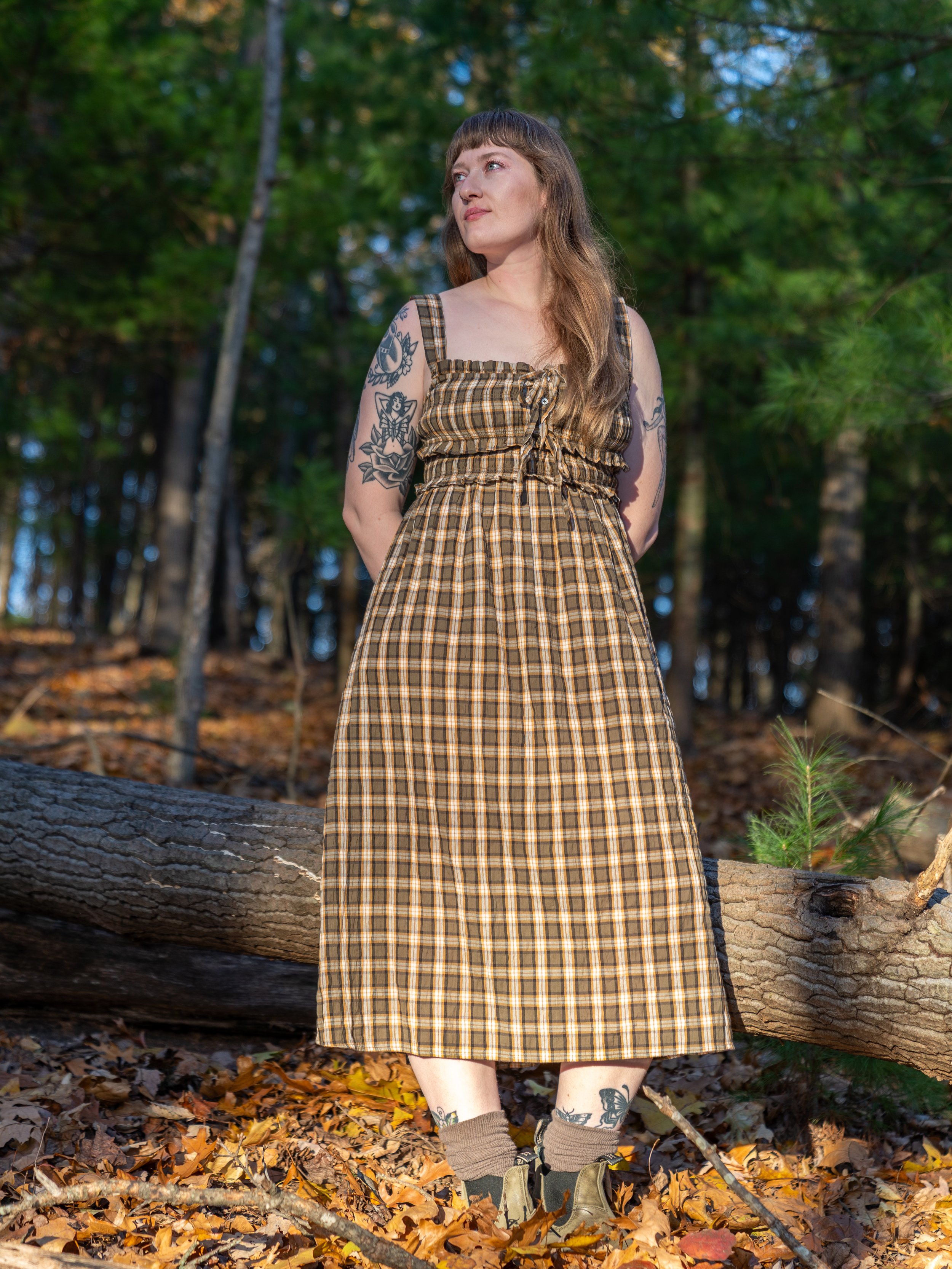 A woman with long brown hair and tattoos on her arms and legs standing outdoors in a forested area on a fall day, wearing a plaid dress, beige socks, and hiking boots.