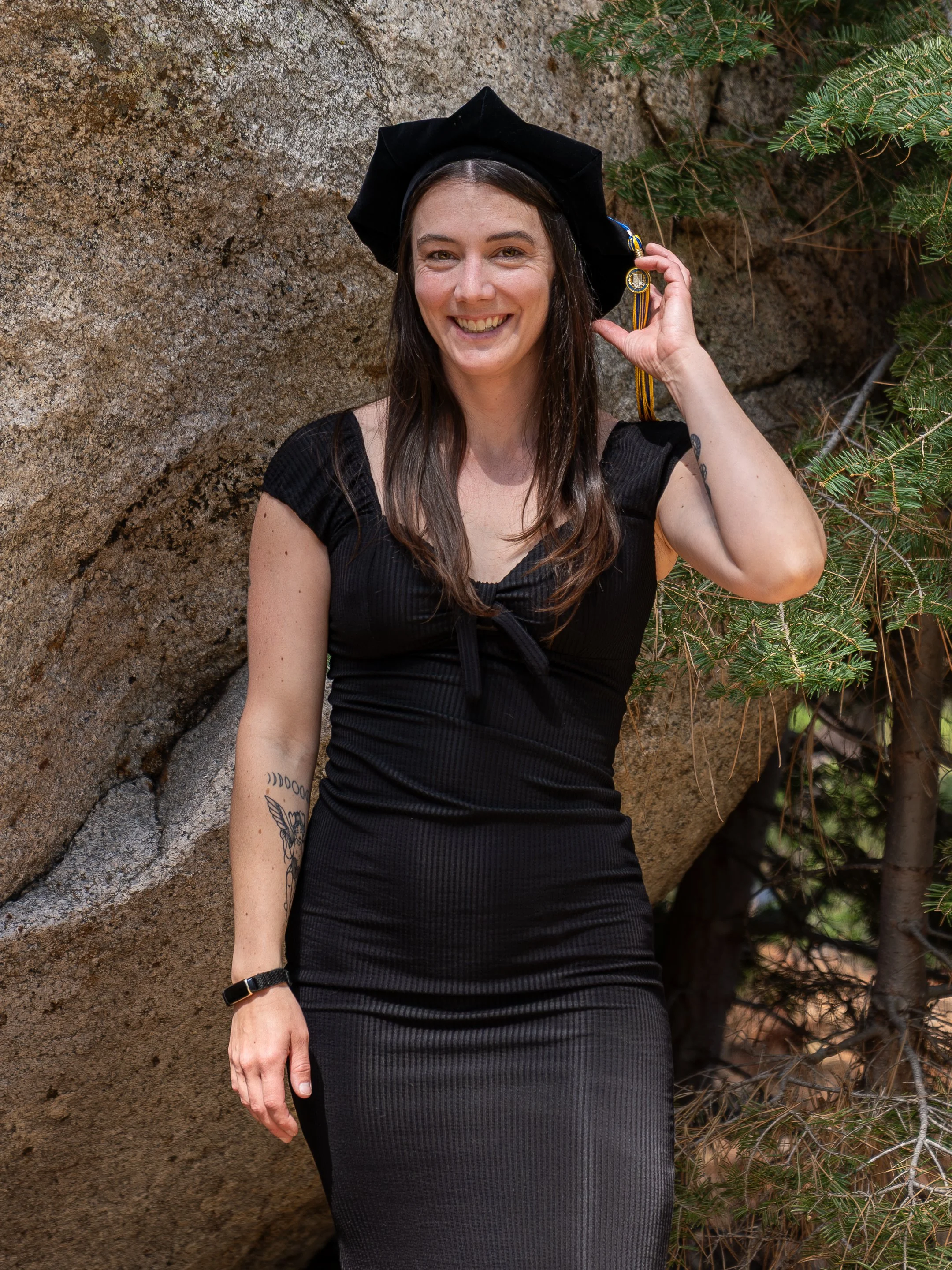 A young woman with tattoos on her arm, wearing a black dress and a black graduation cap, standing outdoors against a rock and pine tree background, smiling and holding her tassel.