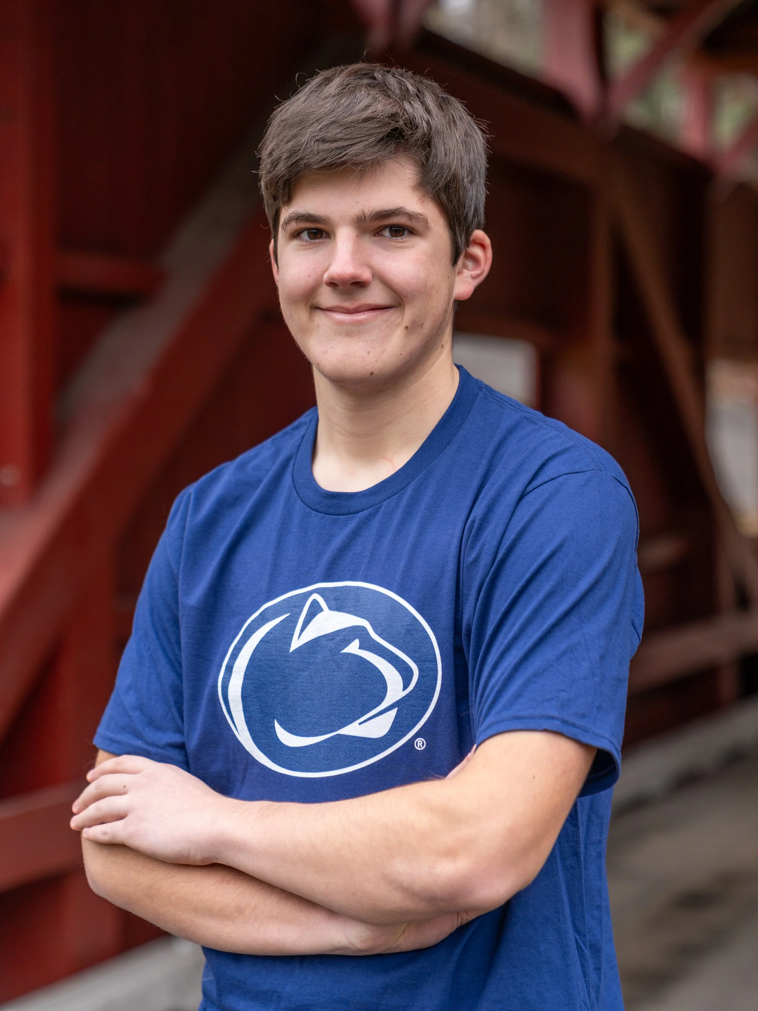 A young man with short brown hair and light skin is smiling with his arms crossed, wearing a blue T-shirt with a white logo on it, in front of a red wooden structure.