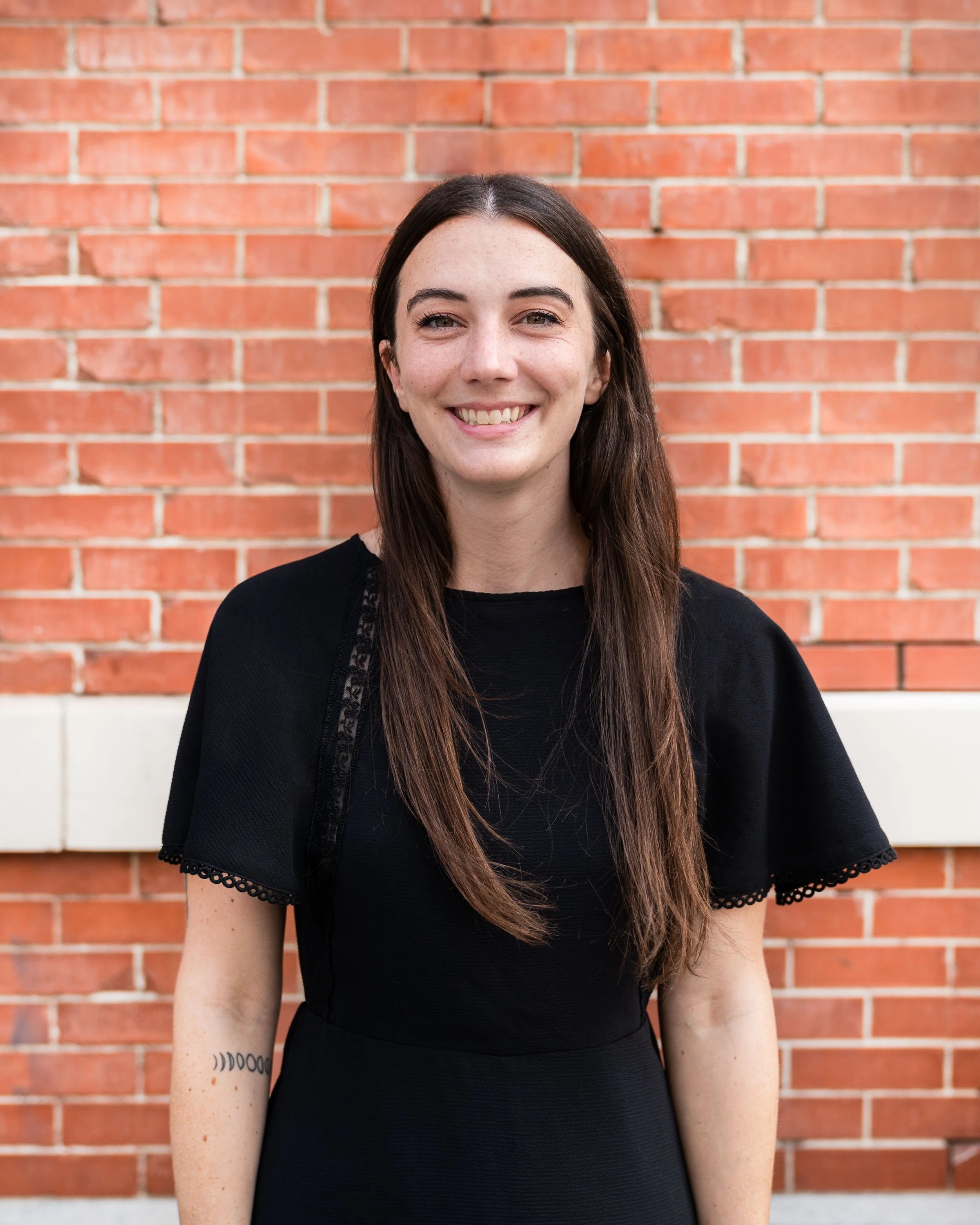A young woman with long brown hair, freckles, and a bright smile, standing in front of a red brick wall. She is wearing a black top with lace detailing on the sleeves.
