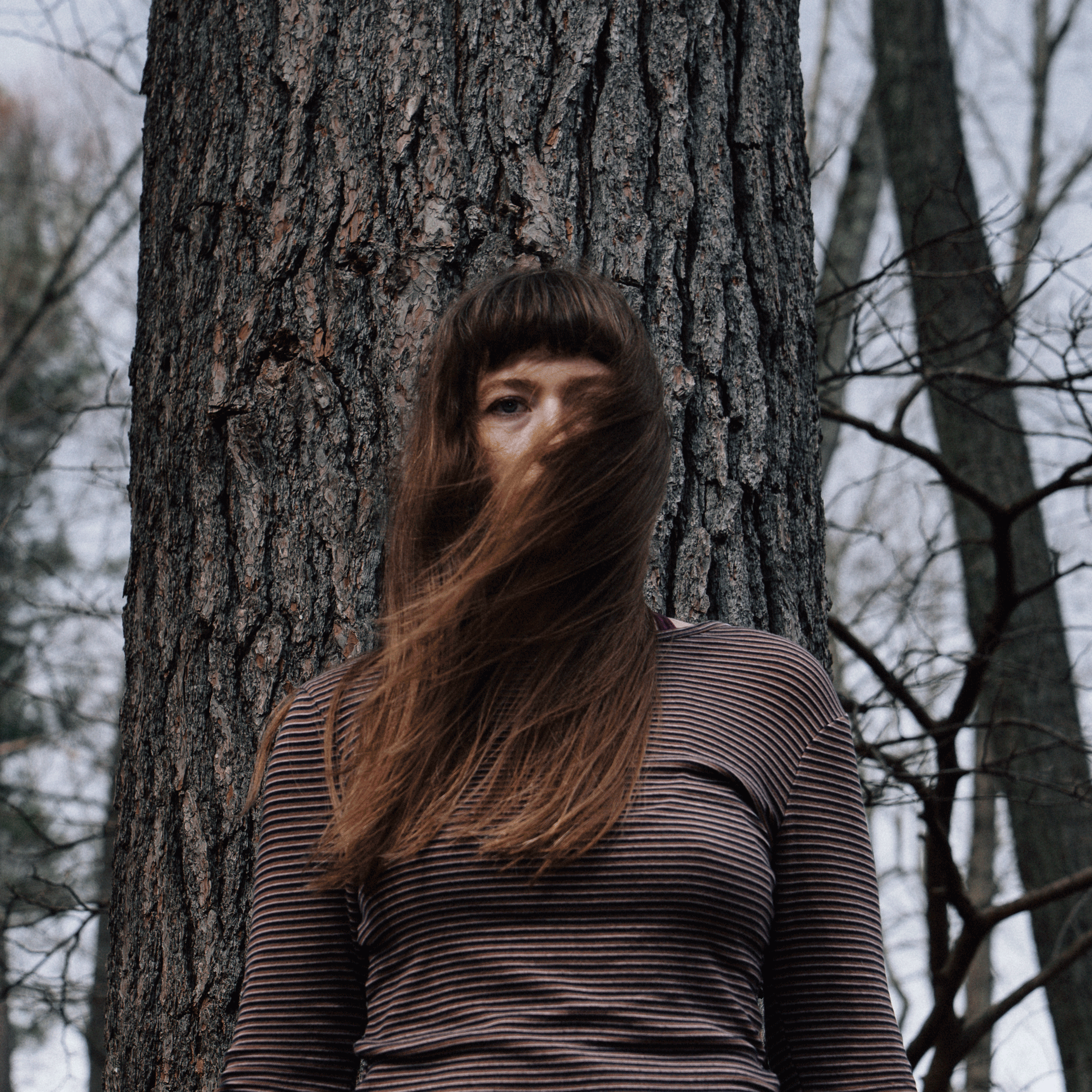 A woman with long hair partially covering her face, standing in front of a large tree trunk in a wooded area during overcast weather.