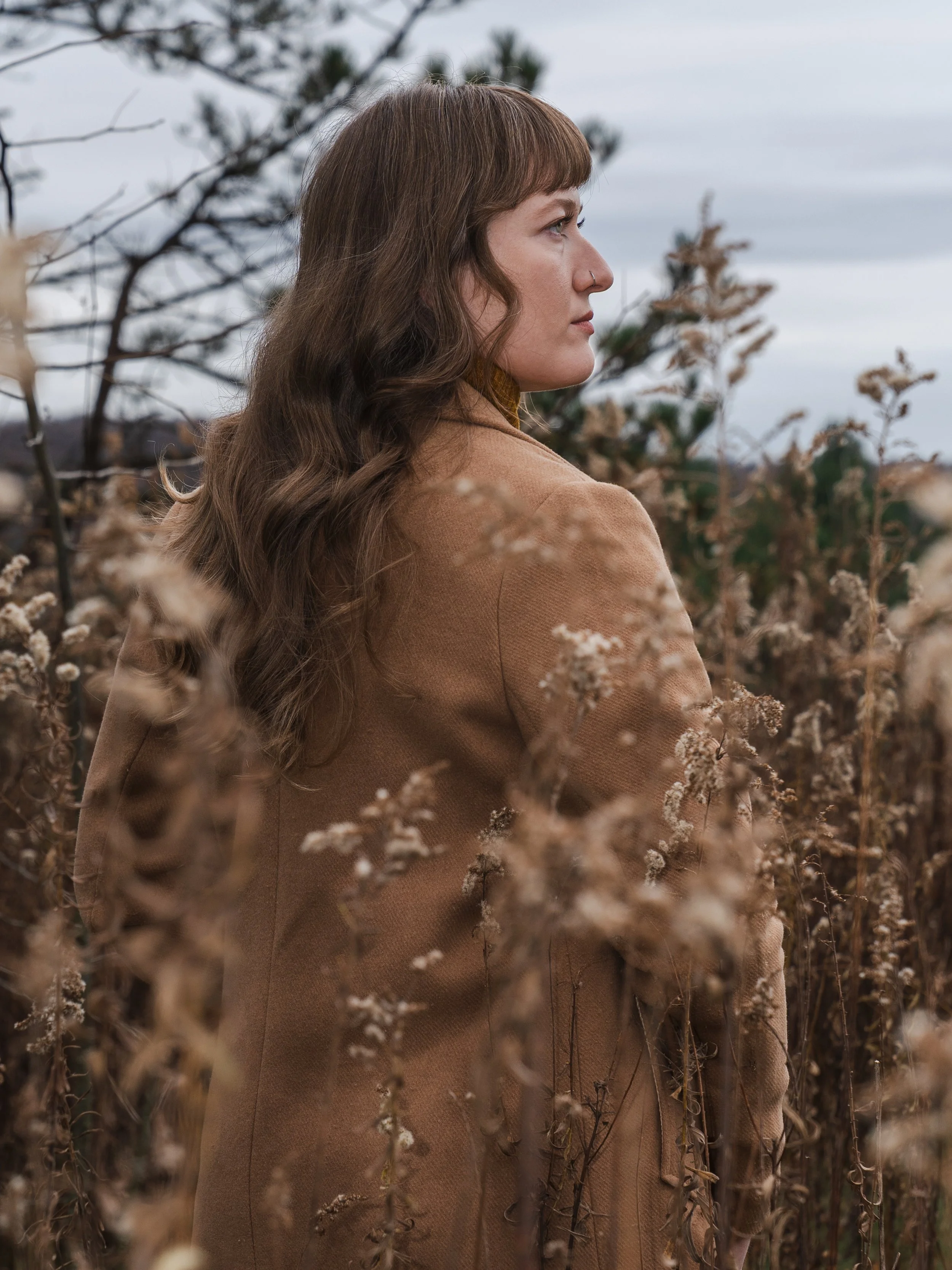 Side view of a woman with brown wavy hair and a nose piercing, wearing a brown coat, standing among dried plants or weeds outdoors on a cloudy day.