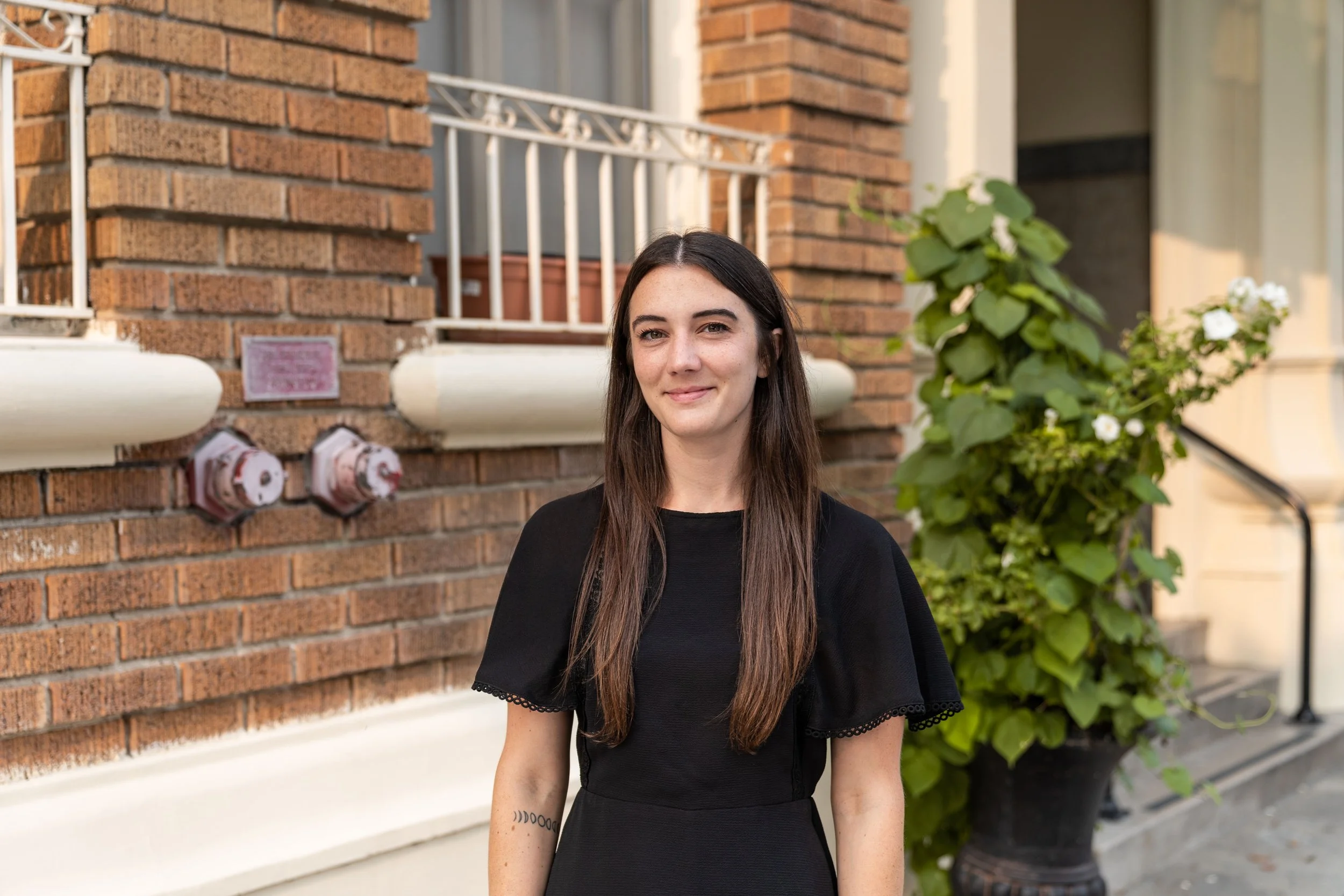 A young woman with long brown hair, smiling, wearing a black top, standing outdoors near a brick building with a white railing, green leafy plants, and a yellow door in the background.