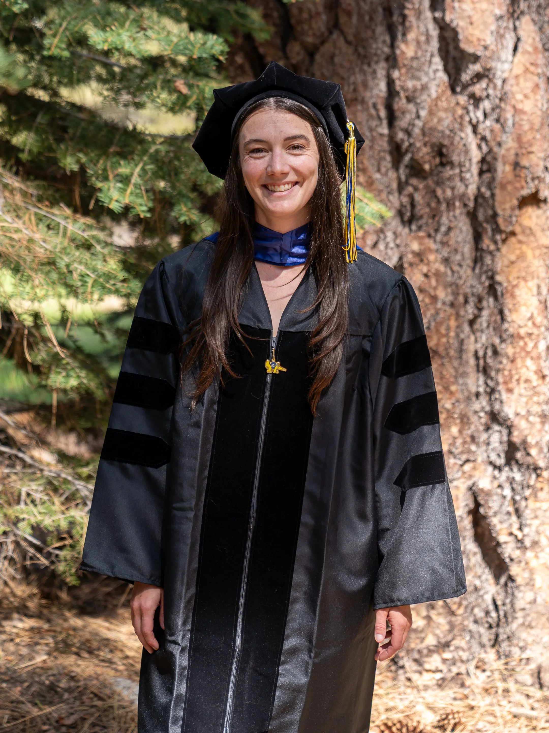 Woman in graduation cap and gown smiling outdoors with trees and a large tree trunk in the background.