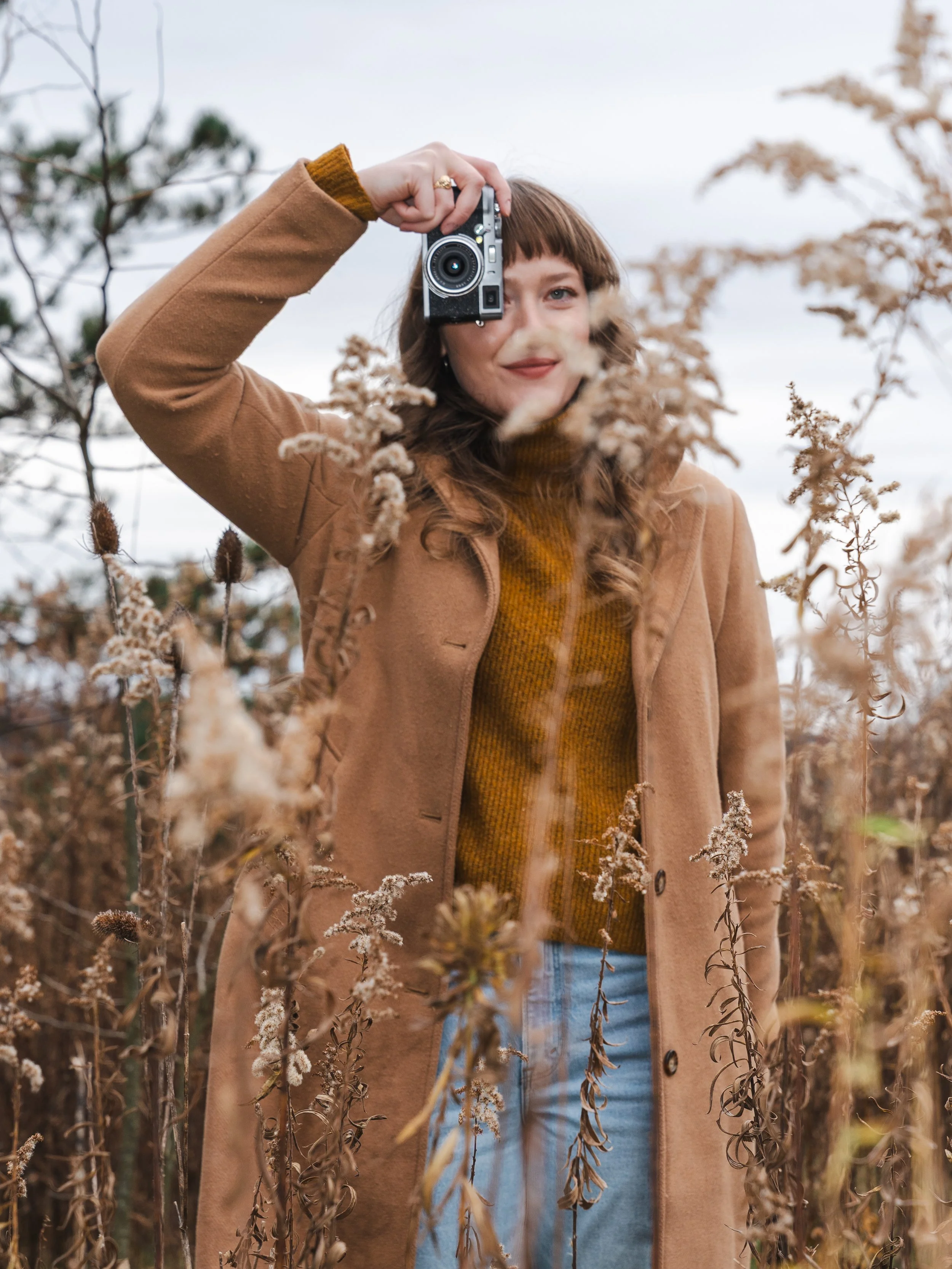 A woman in a brown coat and mustard sweater holding a camera, standing among dry plants outdoors.