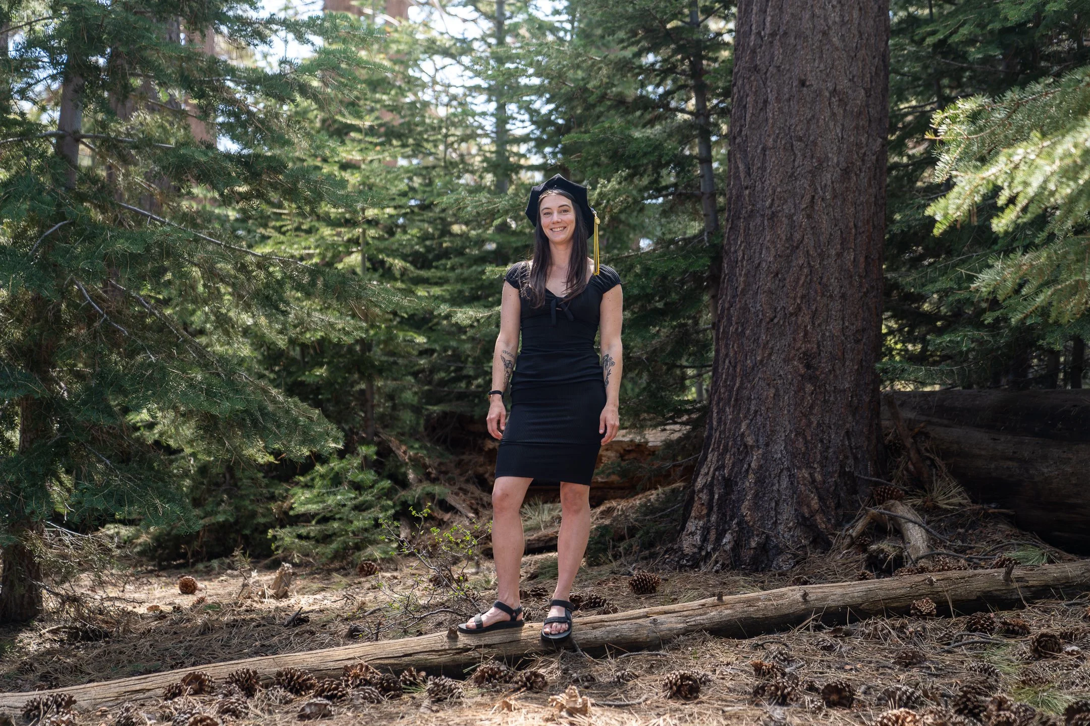 A woman in a black dress and sandals wearing a graduation cap, smiling and standing on a fallen log in a forested area.