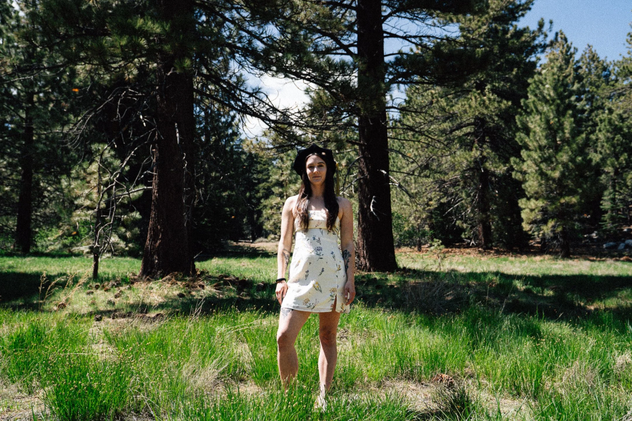 A young woman in a sleeveless dress standing barefoot on grass in a forested area with tall pine trees and blue sky.