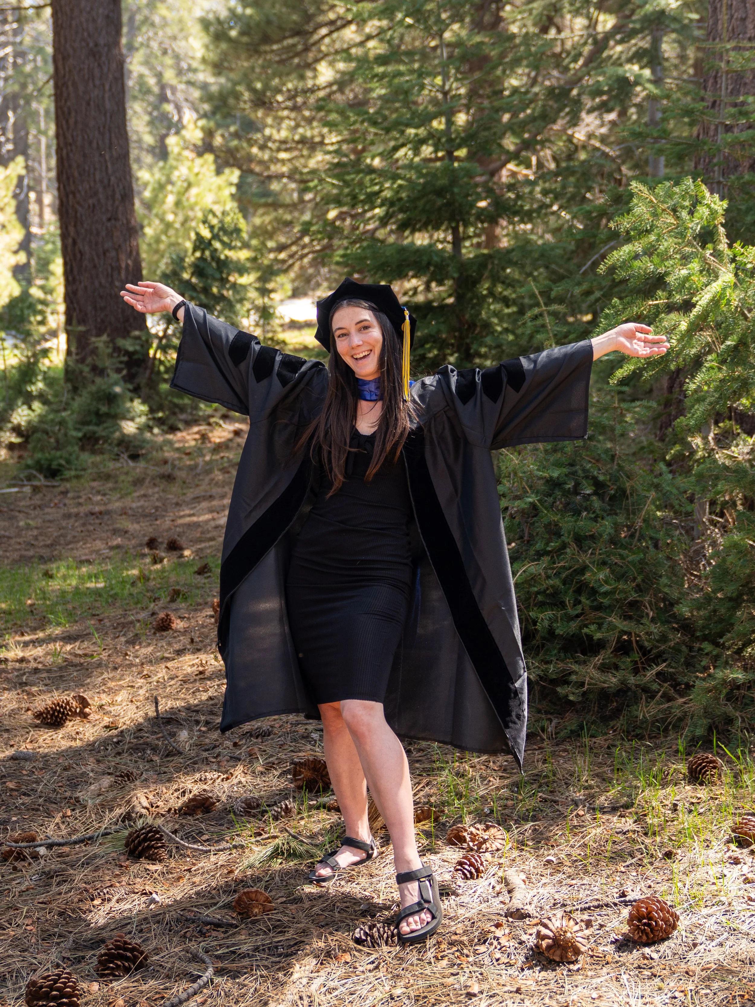 A woman in graduation attire celebrating outdoors in a wooded area with pine cones on the ground.