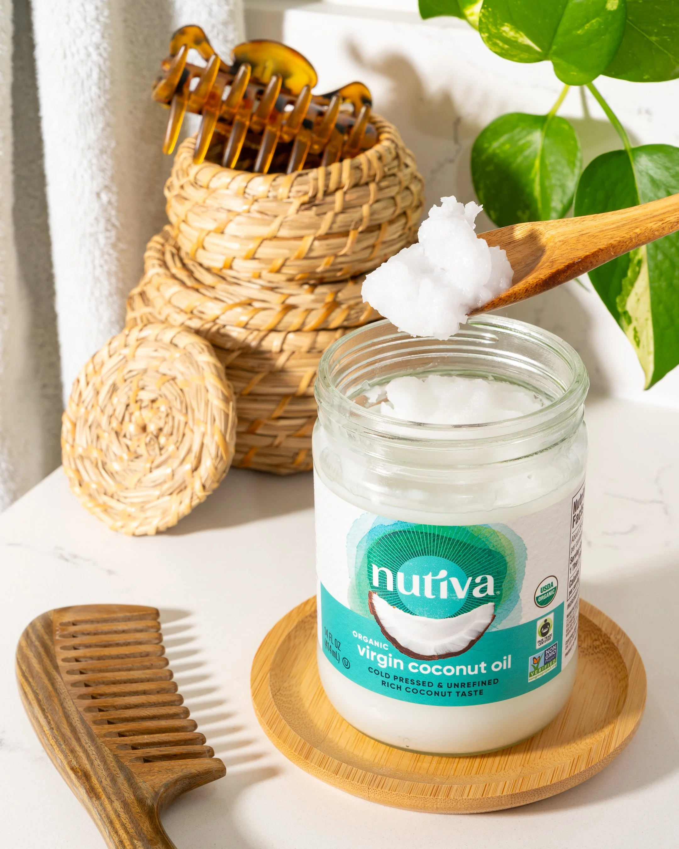 A jar of Nutiva organic virgin coconut oil with a wooden spoon scooping out some. A wooden comb is placed beside the jar on a round wooden tray. In the background, there's a woven basket and a green leafy plant.