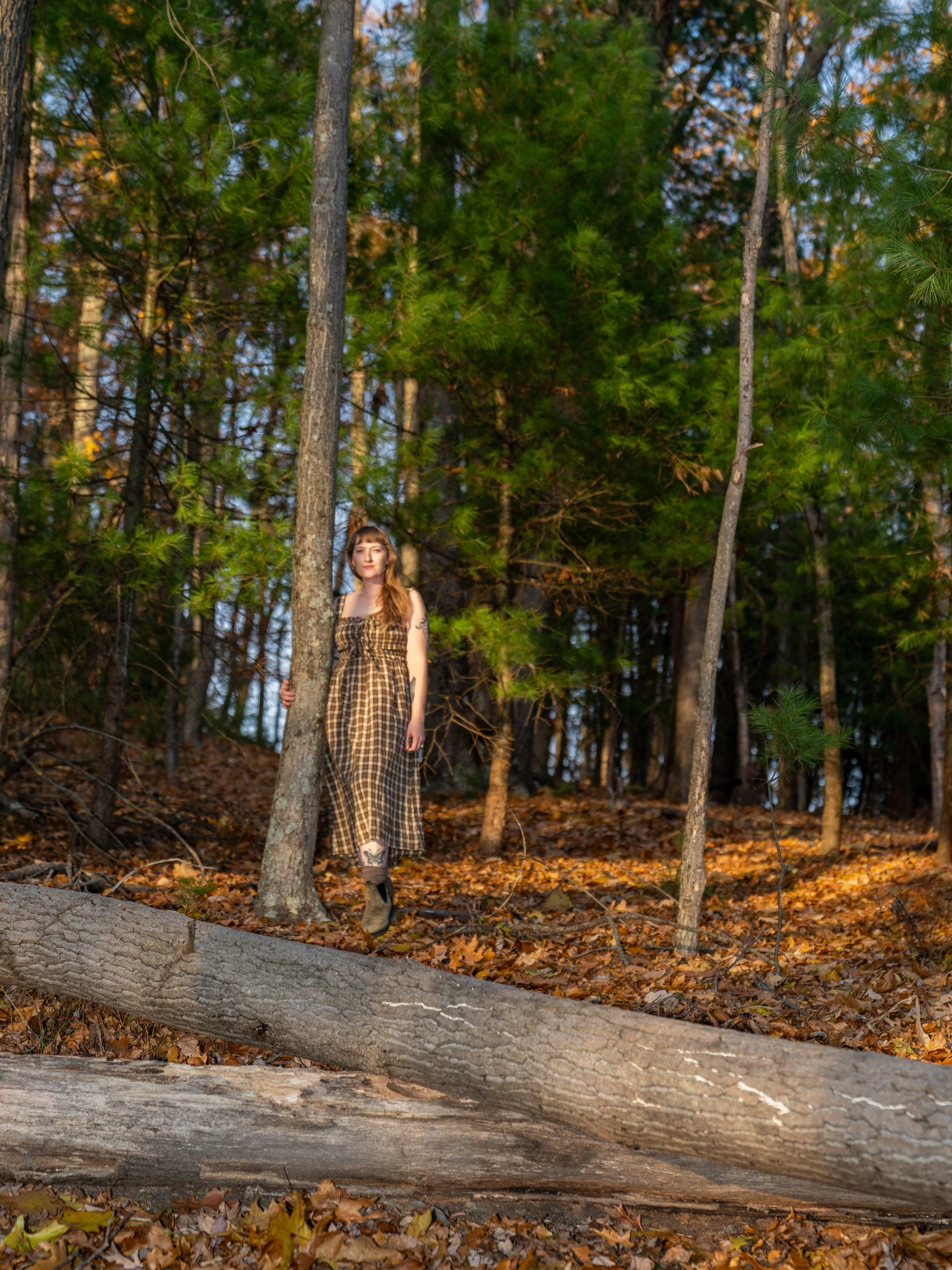 A woman with long reddish-brown hair, wearing a plaid dress and brown boots, standing among trees and fallen autumn leaves in a forested area.