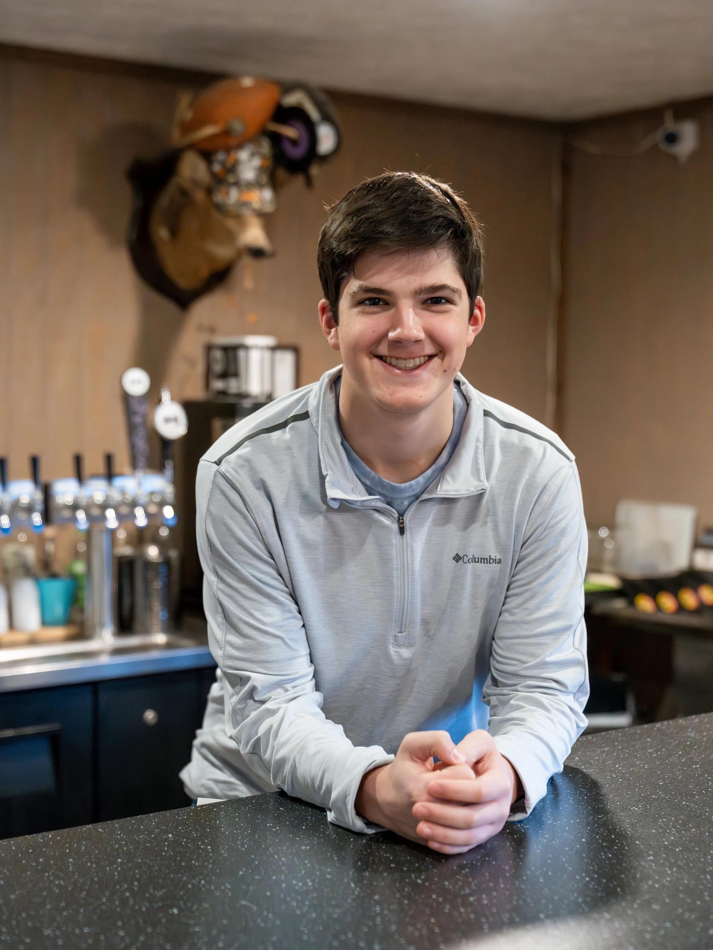 A young man with short dark hair smiling, leaning on a speckled black counter in a casual kitchen or bar area, wearing a light gray Columbia jacket. In the background, there is a wooden mounted animal head and beer taps on the counter.