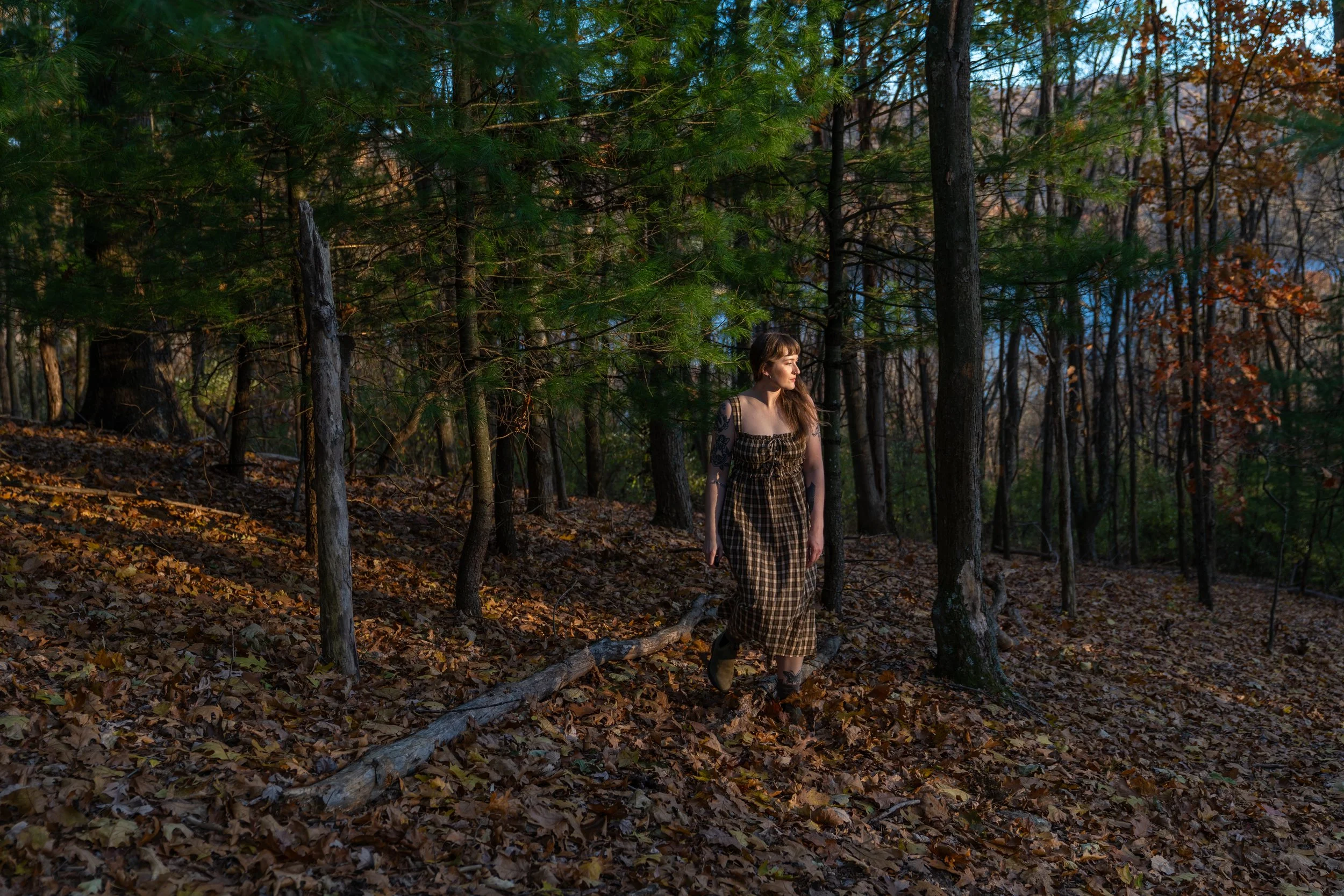 A young woman with long brown hair, wearing a checkered dress, walking through a wooded forest area with fallen leaves on the ground during late afternoon or early evening.