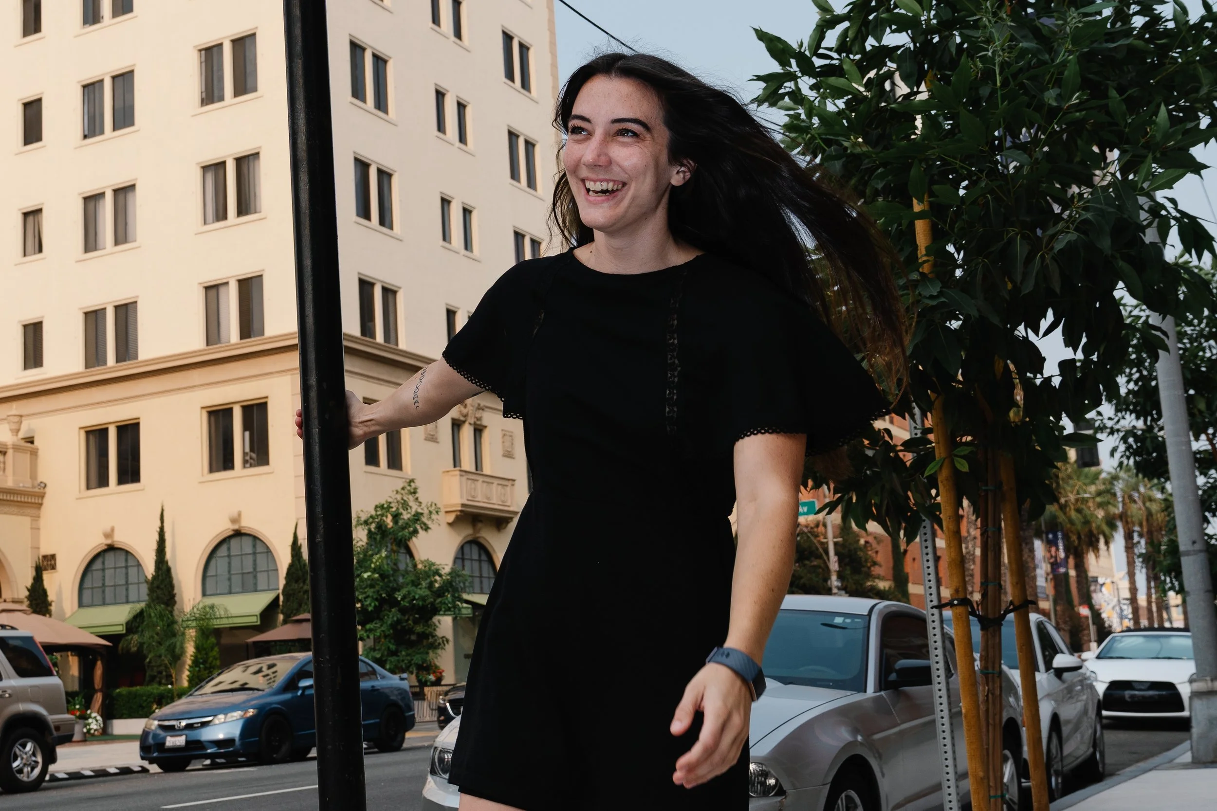 A woman with long dark hair smiling and walking outdoors in an urban area with cars parked on the street, trees, a black pole, and a tall beige building with multiple windows in the background.