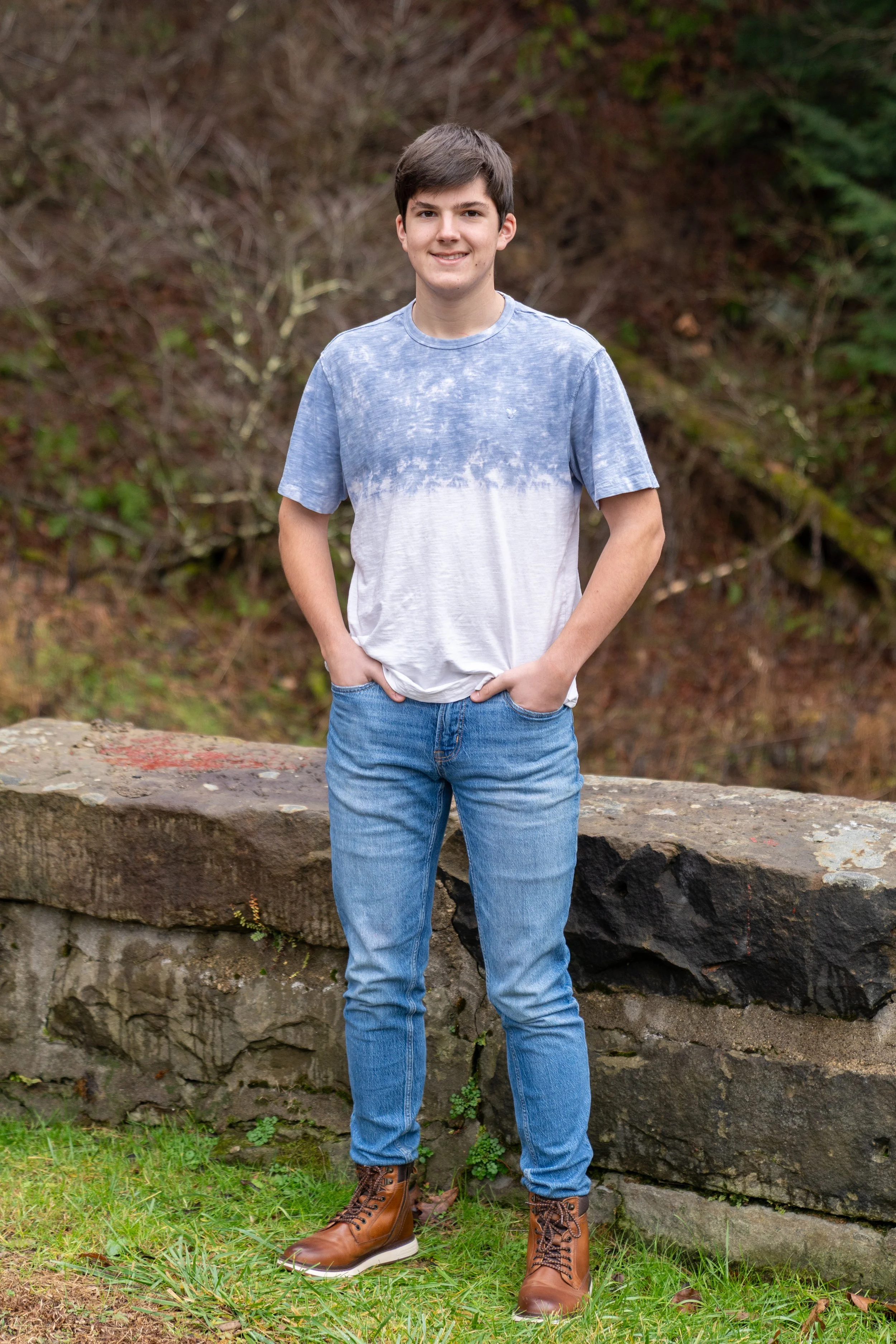 A young man with dark hair, wearing a blue and white tie-dye t-shirt, blue jeans, and brown boots, standing outdoors in front of a stone wall and foliage, smiling at the camera.