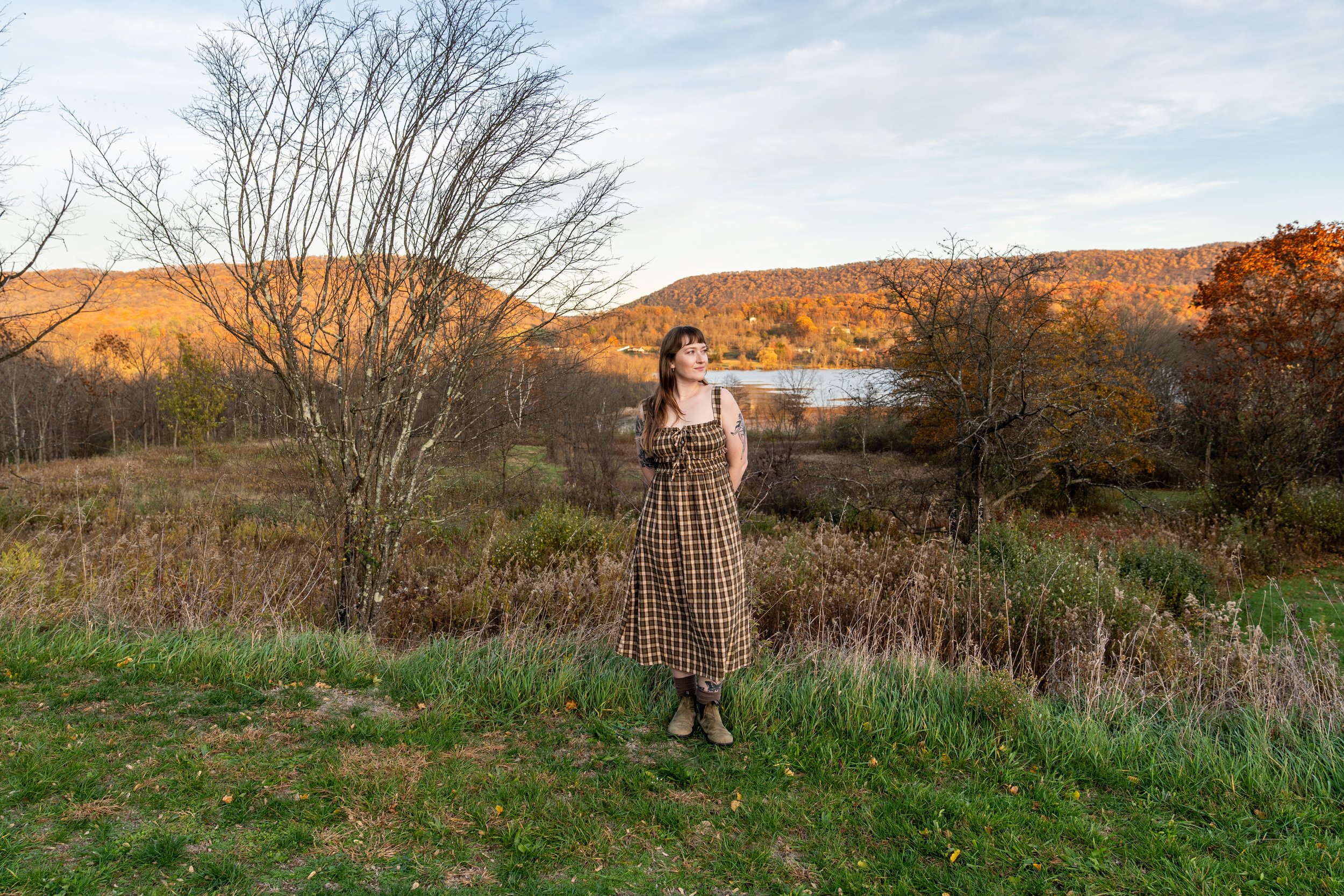 A woman with brown hair and a checkered dress stands outdoors near a grassy area with autumn trees and hills in the background. A body of water can be seen in the distance under a partly cloudy sky.