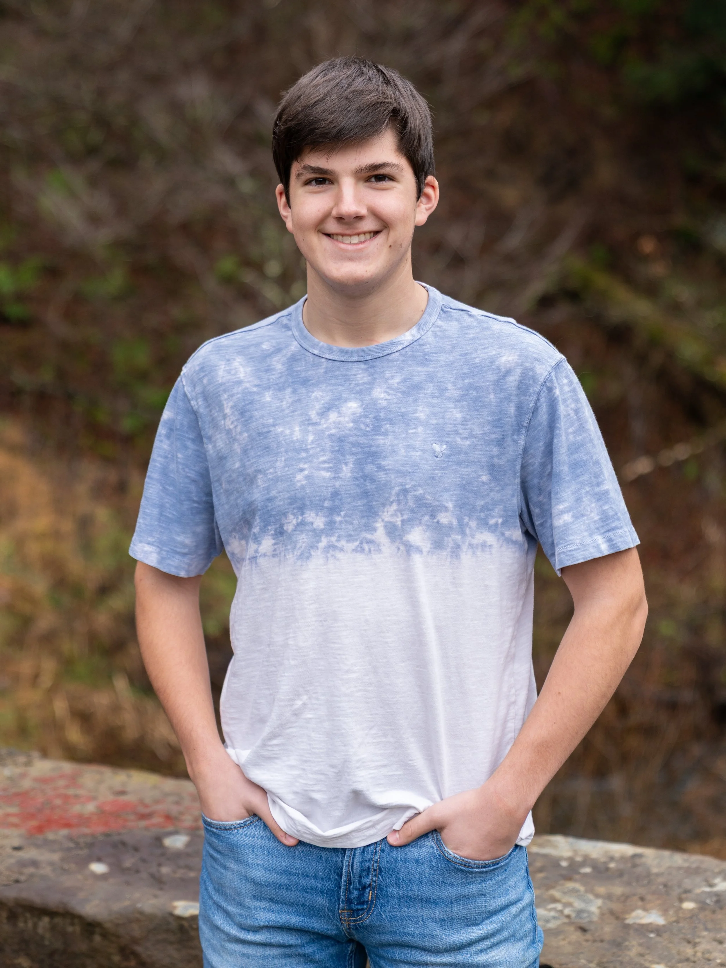 A young man with dark hair smiling, wearing a blue and white tie-dye T-shirt and blue jeans, standing outdoors in front of a natural background.