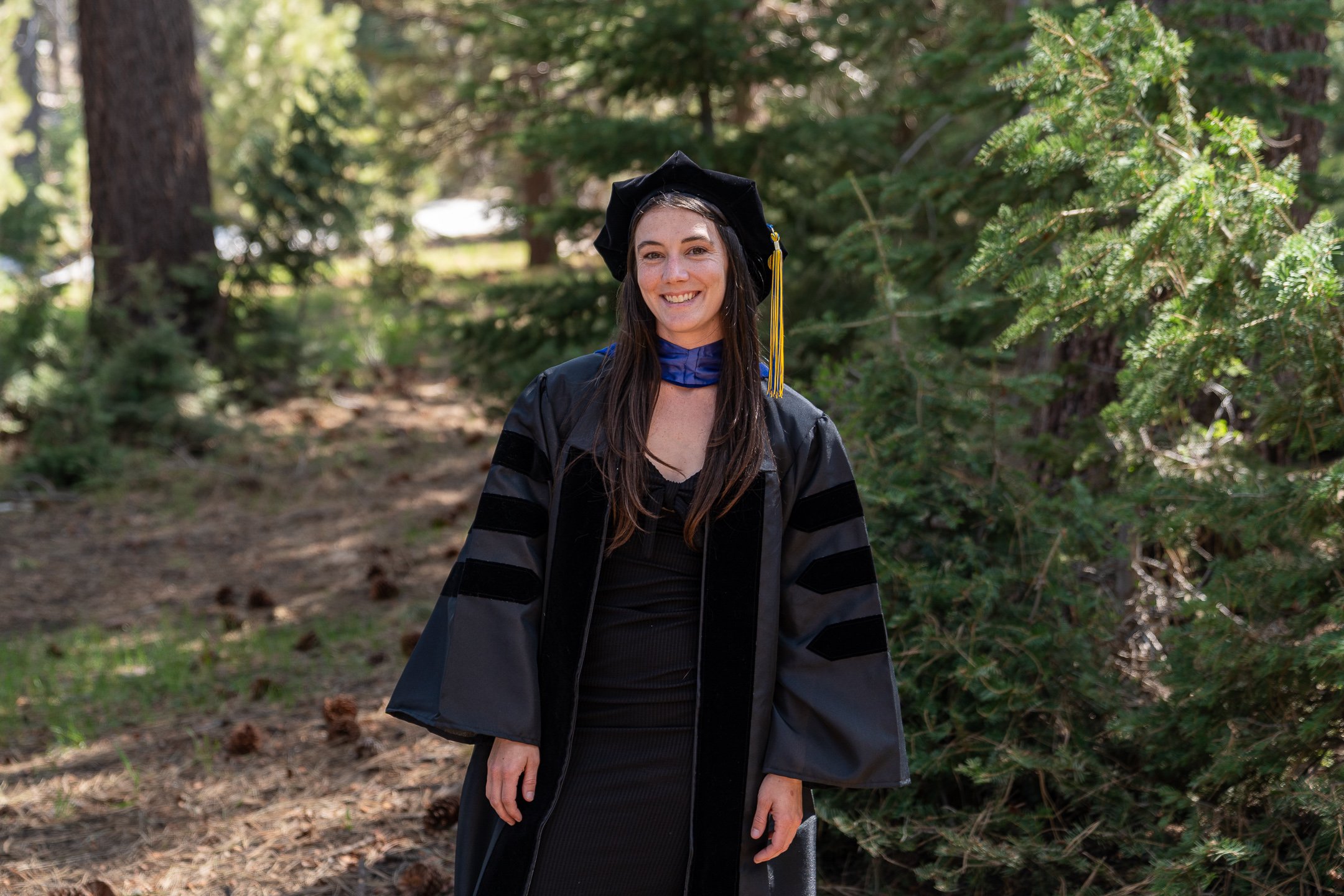 A woman in graduation attire standing outdoors in a wooded area, wearing a black gown, a black cap with a yellow tassel, and a blue stole, smiling at the camera.