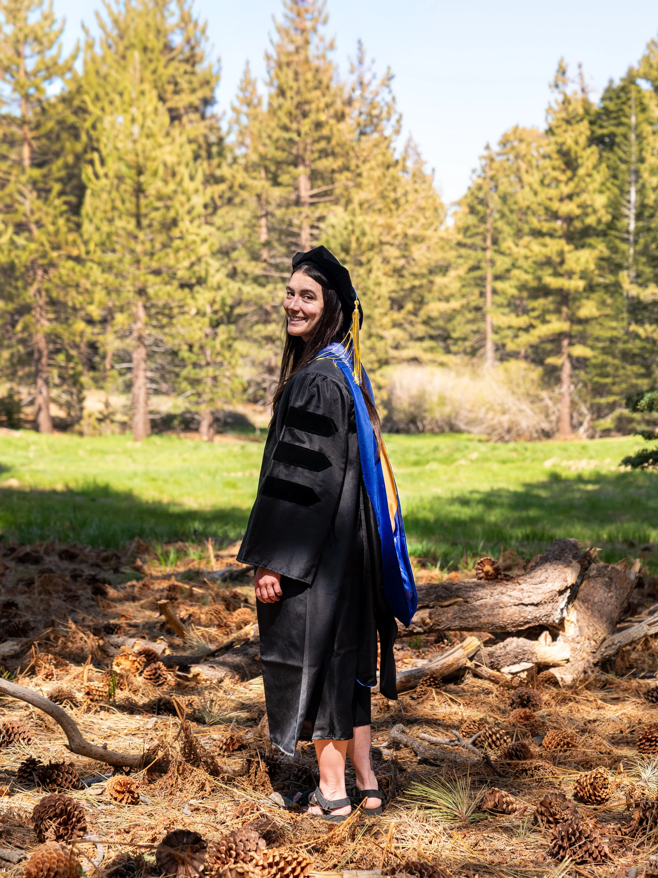A woman in graduation cap and gown standing outdoors on a forest trail with pine trees and fallen pinecones.