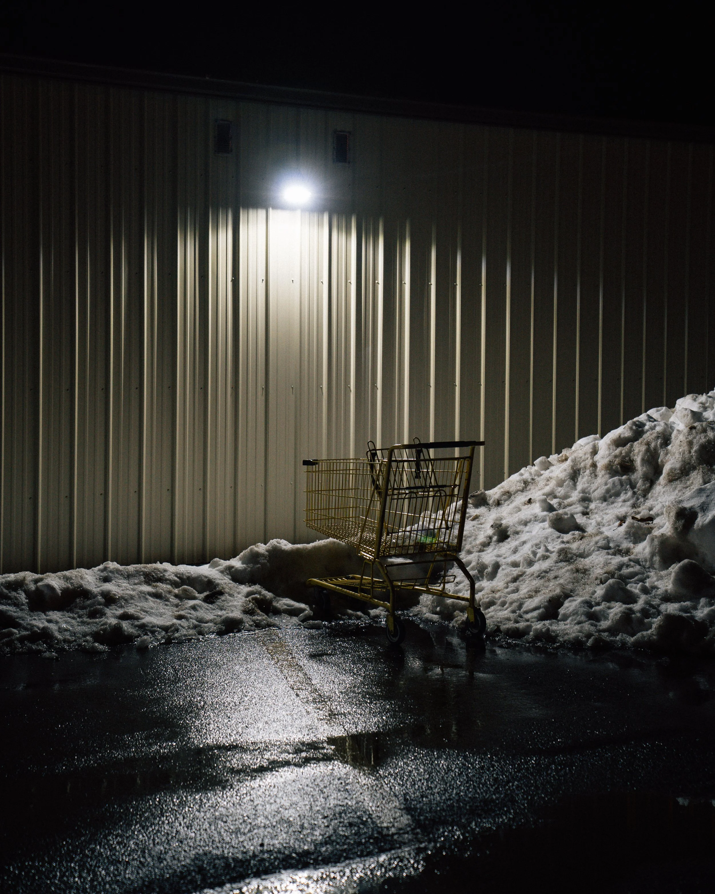 An empty shopping cart on wet pavement outside a building at night, with snow piled against the wall, illuminated by a bright exterior light.