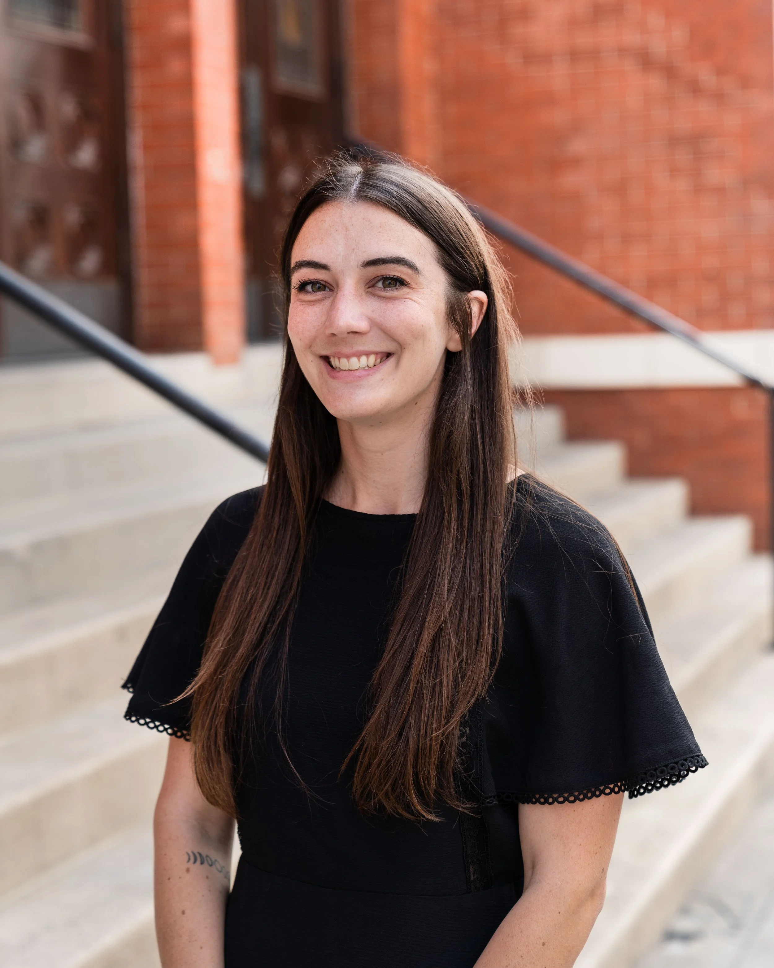 Close-up of a smiling woman with long brown hair, wearing a black top, standing outdoors in front of a brick building with stairs.