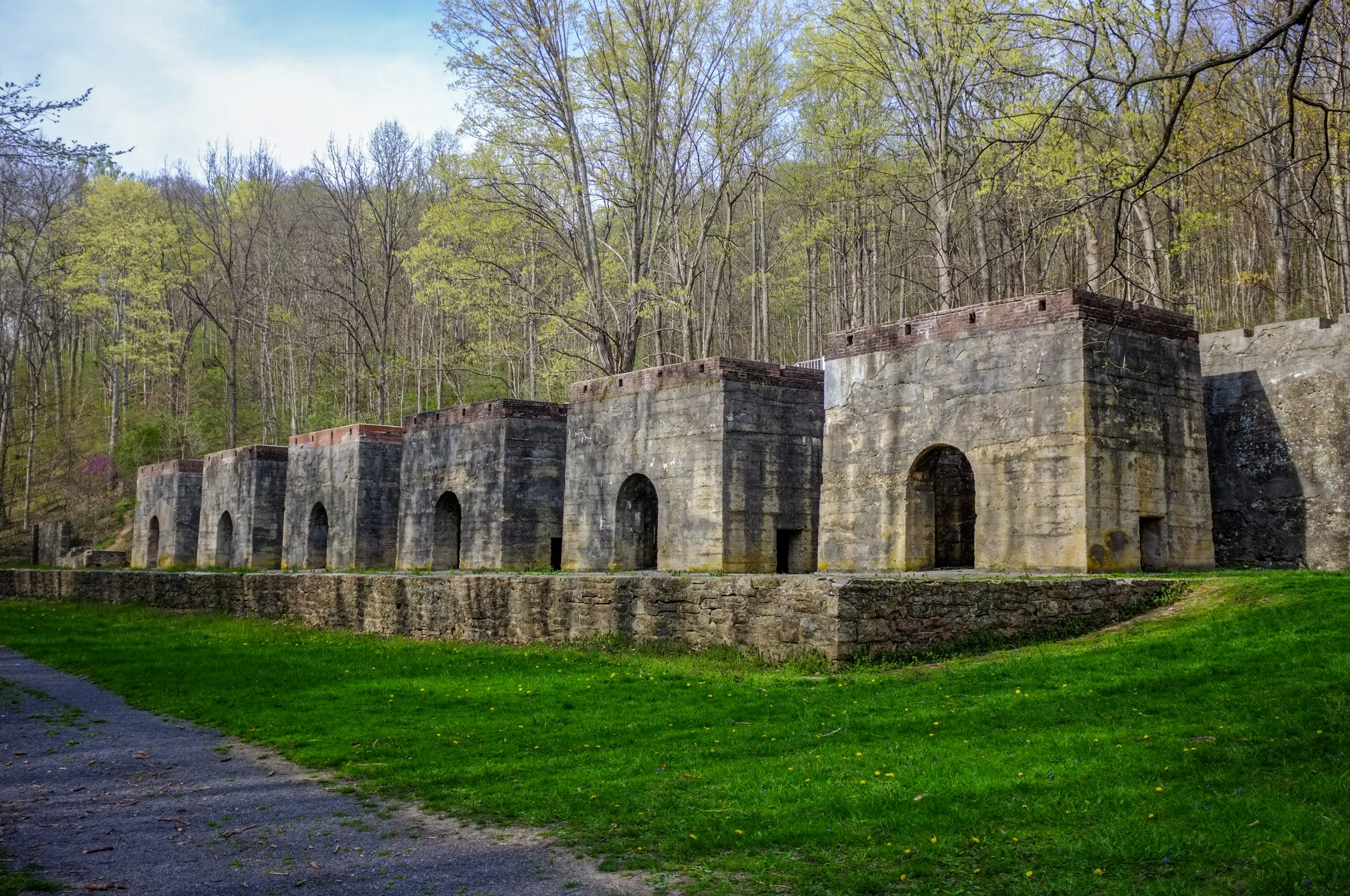 Historic stone and brick structures with arched openings, set on a grassy area at the base of a forested hillside.