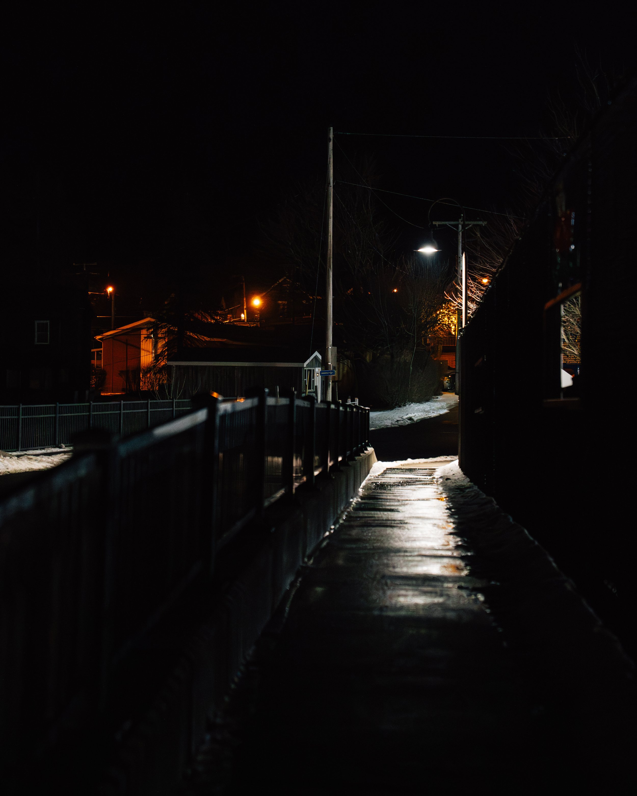 Nighttime scene of a narrow, wet sidewalk with a metal railing, nearby houses with lights, power poles, and snow on the ground.