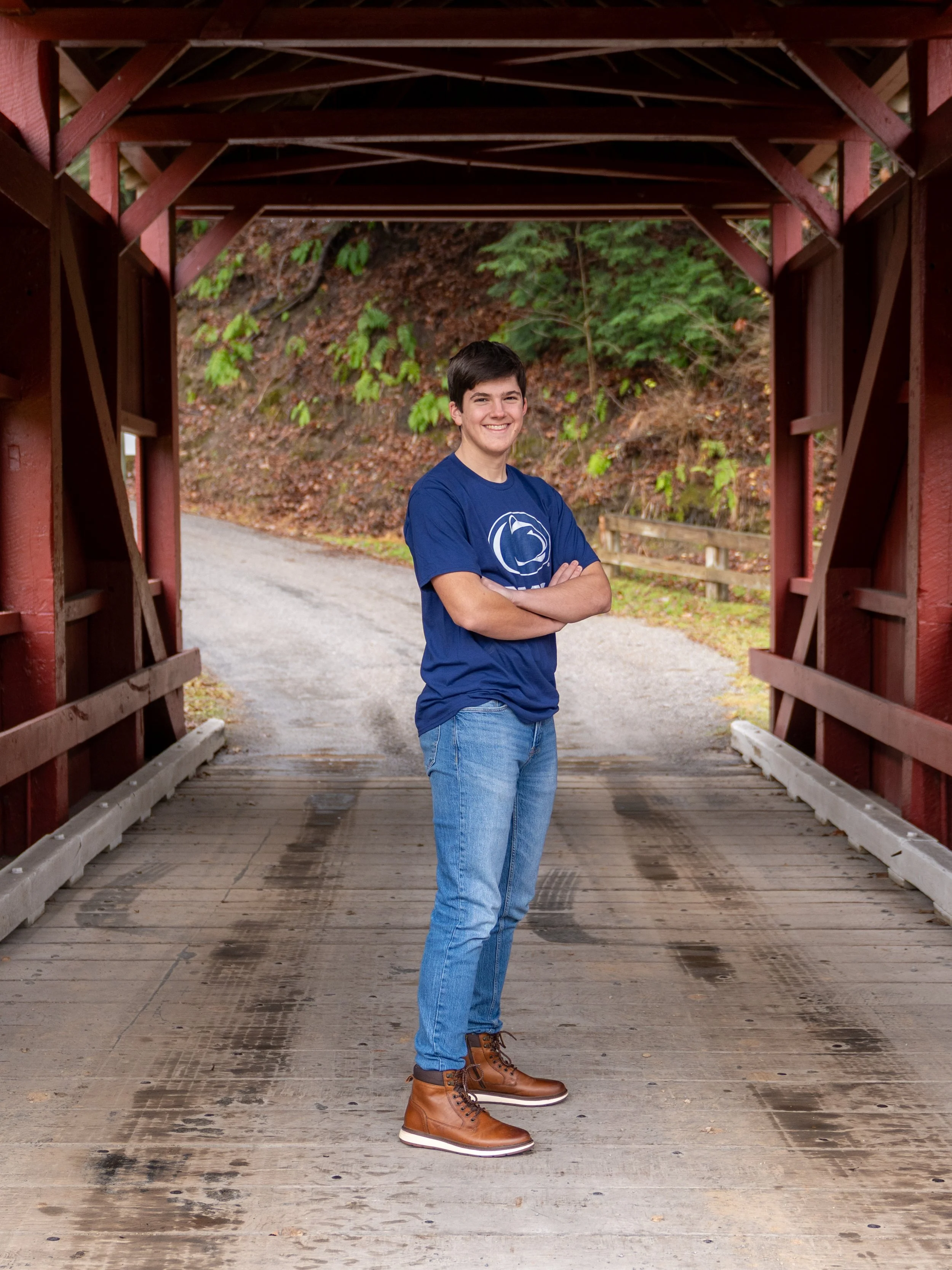 A young man with dark hair and a smile, standing with arms crossed on a covered wooden bridge outdoors, wearing a blue t-shirt, light blue jeans, and brown boots.