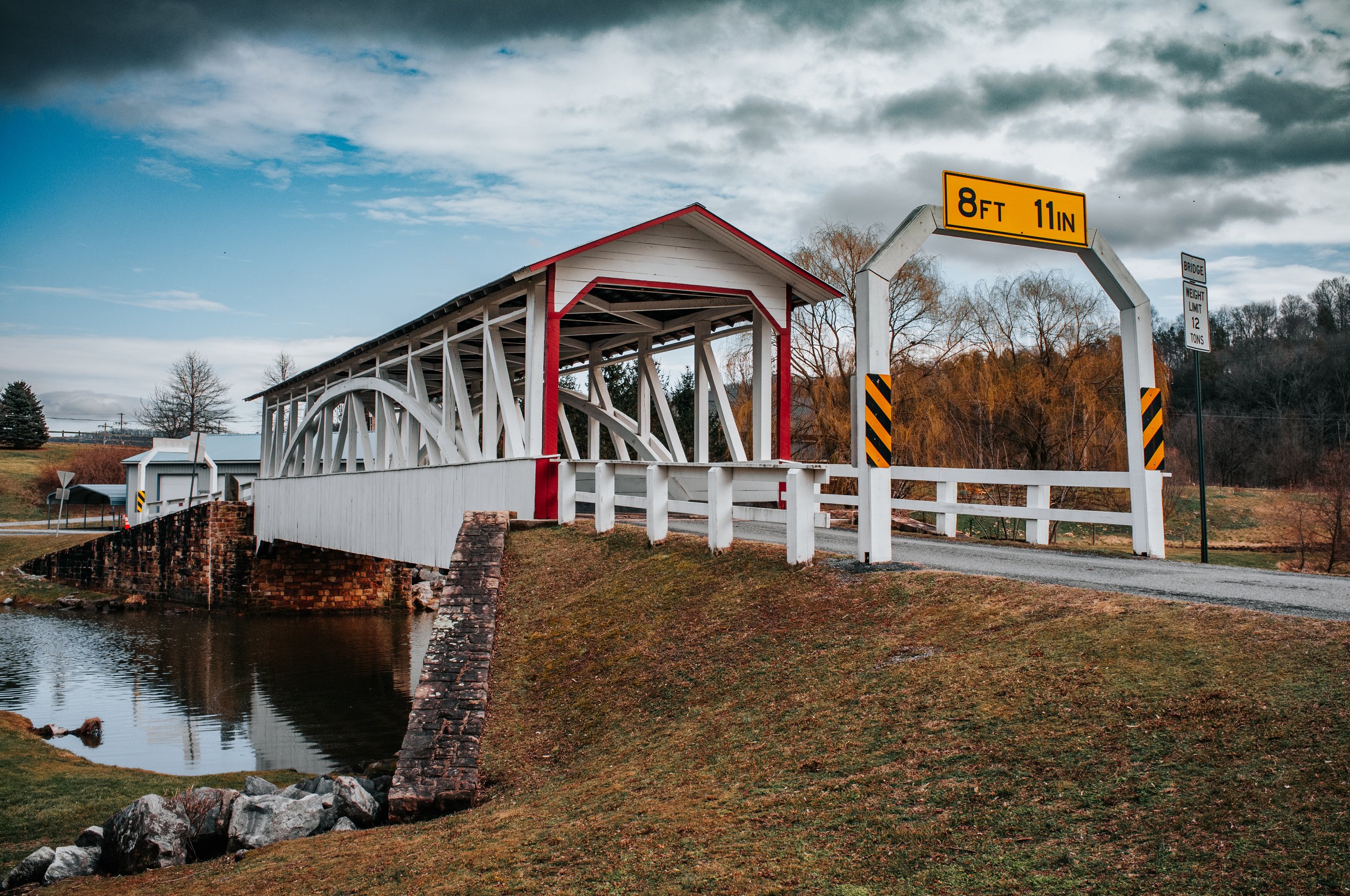 A white covered bridge with red trim spans a small creek in a rural area on a cloudy day. A yellow clearance height sign indicates 8 feet 11 inches. A black and white bridge weight limit sign specifies 12 tons.