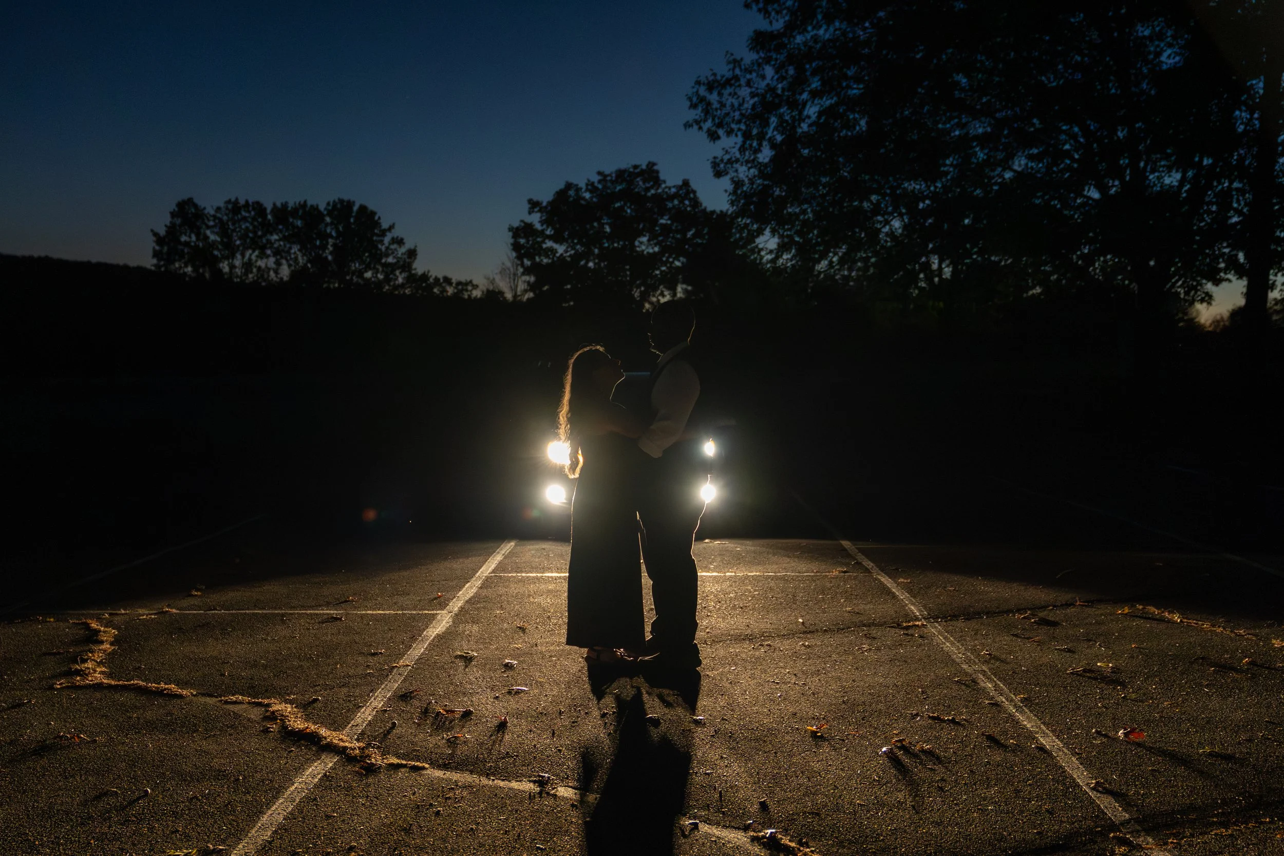 A silhouette of a couple standing close together in a parking lot at night, with their faces near each other, illuminated by the headlights of a car behind them. The sky is dark with some trees visible in the background.