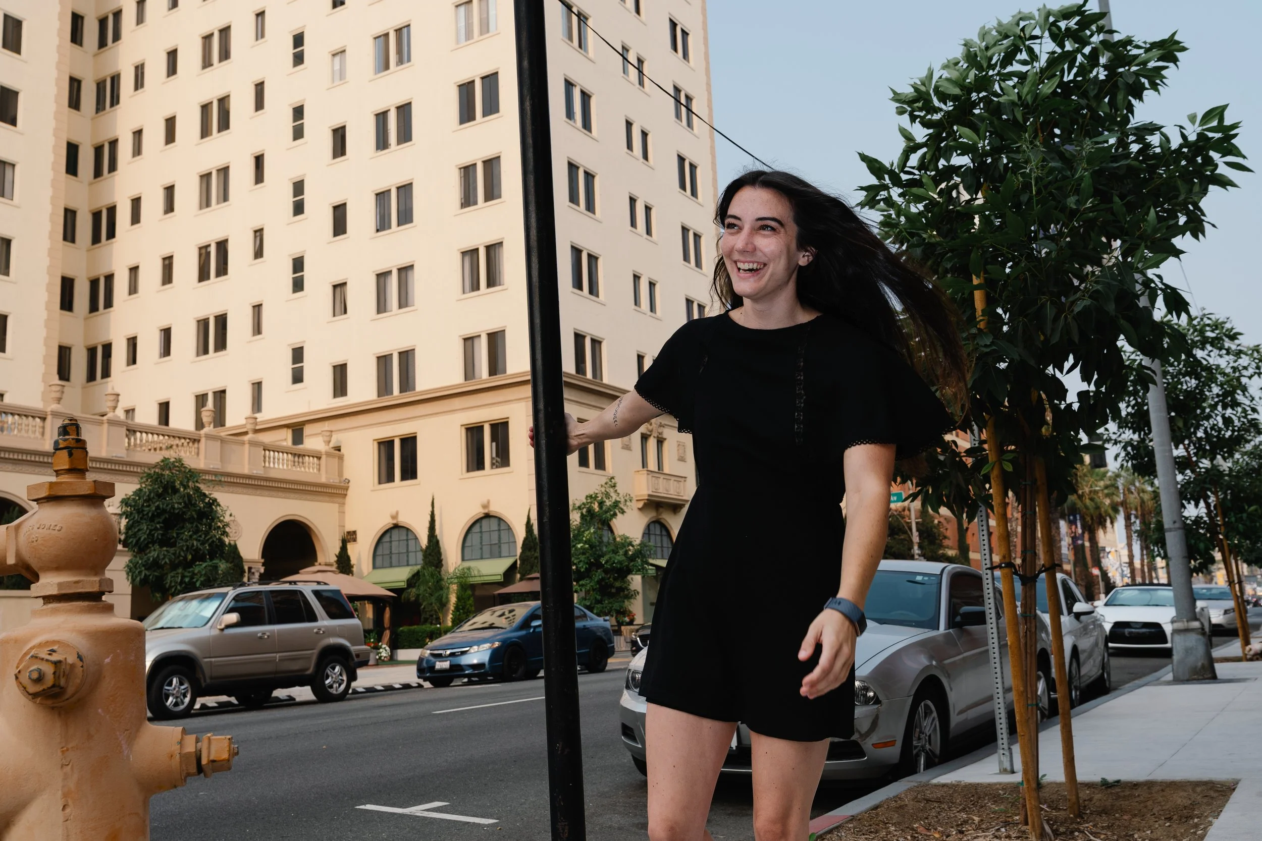 A woman with dark hair in a black dress smiling while holding onto a pole on a city street. There are parked cars and a tall building in the background, along with trees and a fire hydrant in the foreground.