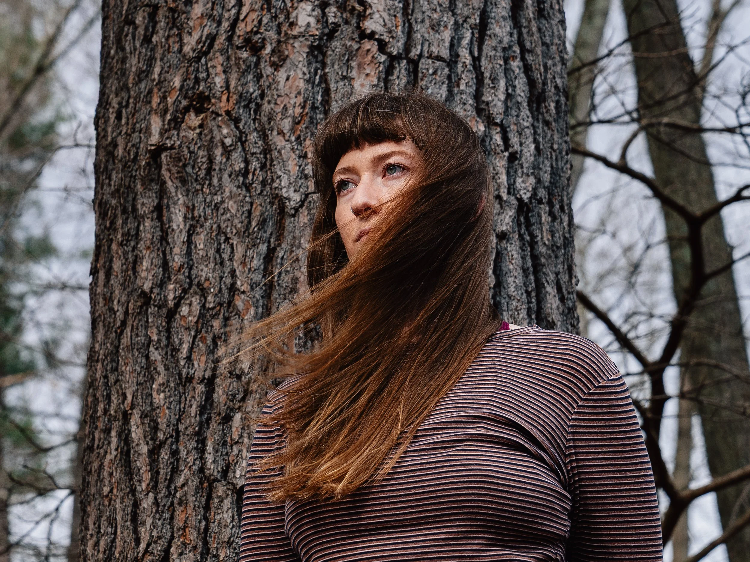 A woman with long brown hair and bangs leans against a large tree trunk outdoors, looking off to the side with a serious expression.