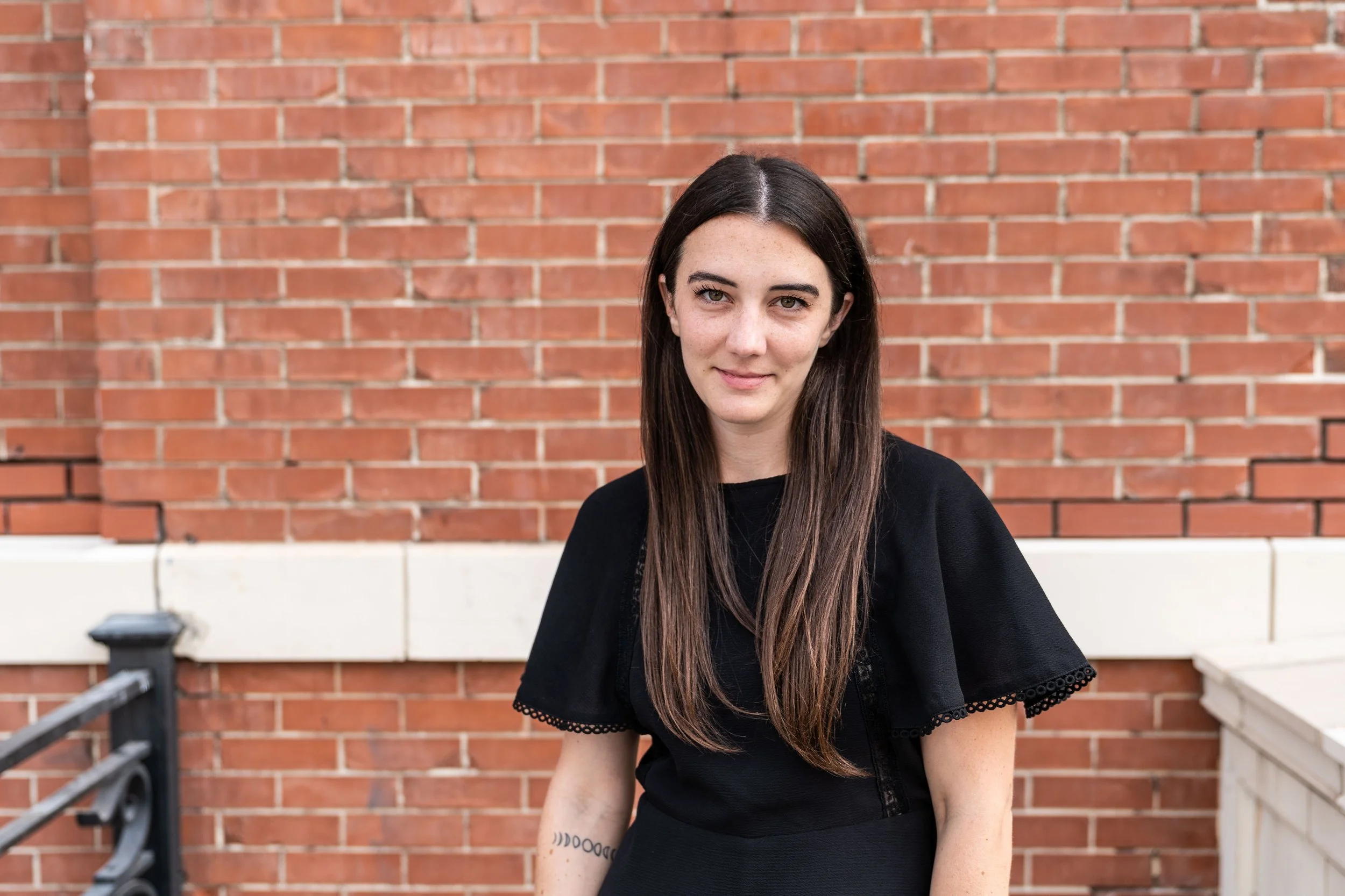A young woman with long dark hair and freckles, wearing a black top, sitting outdoors against a red brick wall.