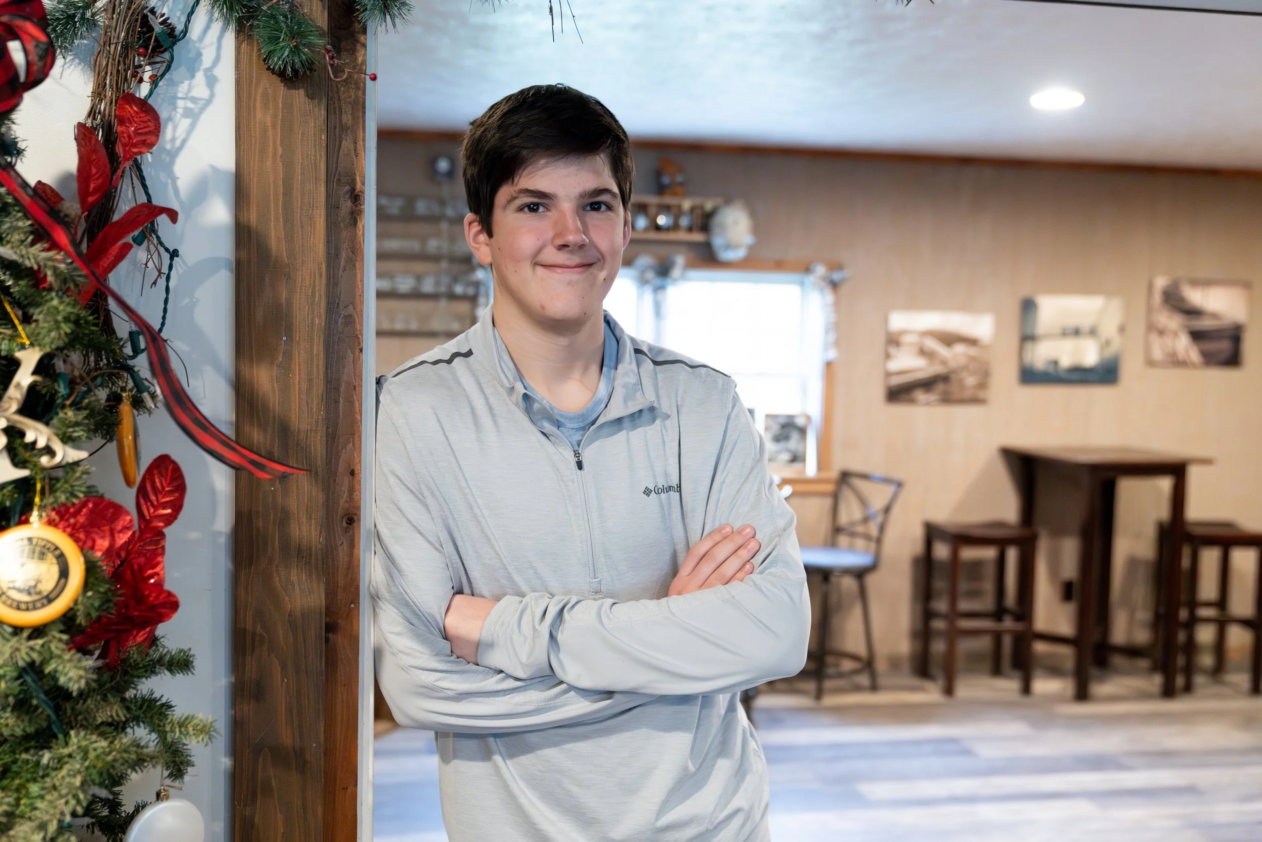 A teenage boy standing indoors near a decorated Christmas tree and a wooden post, with arms crossed, smiling slightly, wearing a light gray Columbia jacket. The background features windows, wall art, and a dining area.