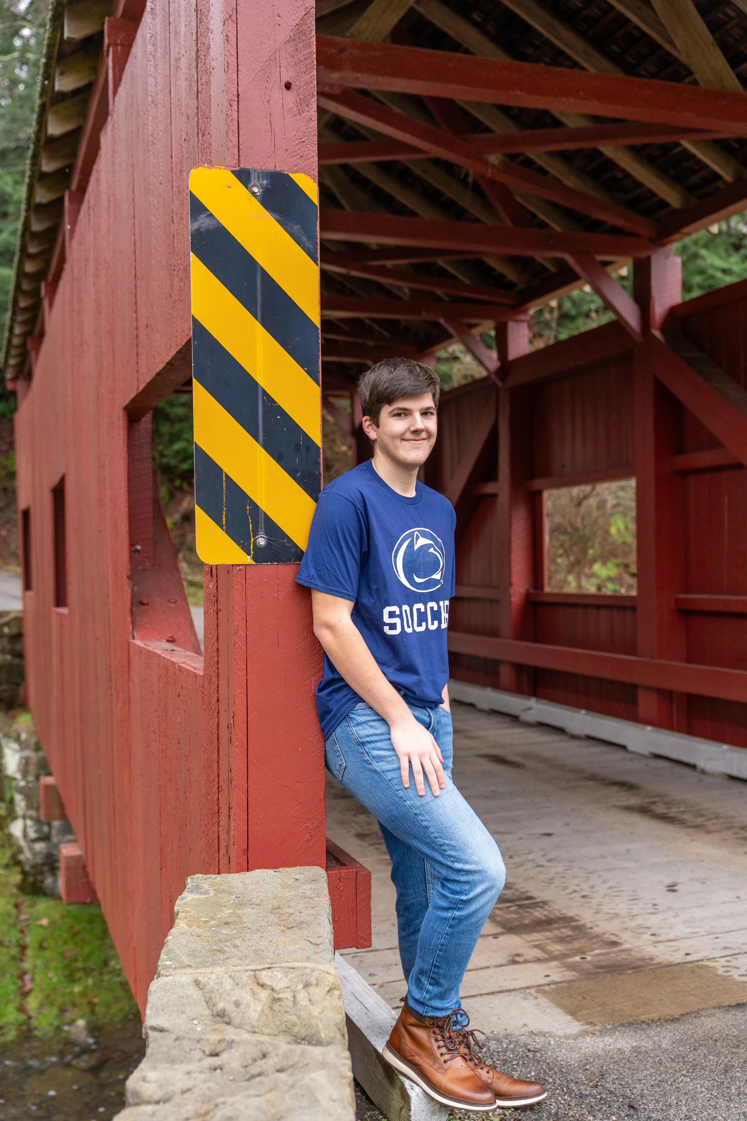 A young man standing under a red wooden covered bridge, leaning against a support post, wearing a blue "Socco" t-shirt, jeans, and brown leather boots.