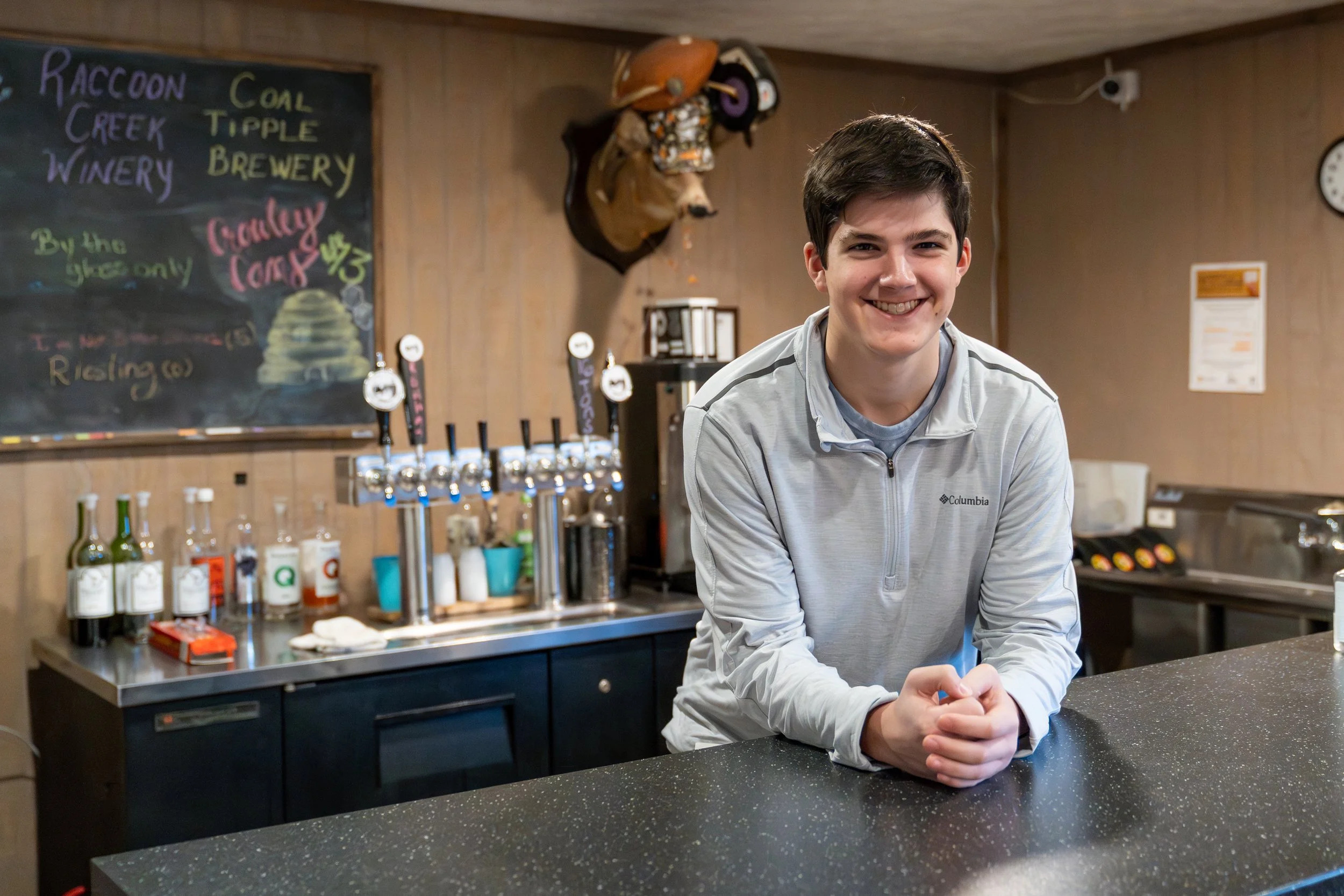A young man smiling at the camera, standing behind a bar counter in a casual bar or brewery setting, with a chalkboard menu and beer taps in the background.