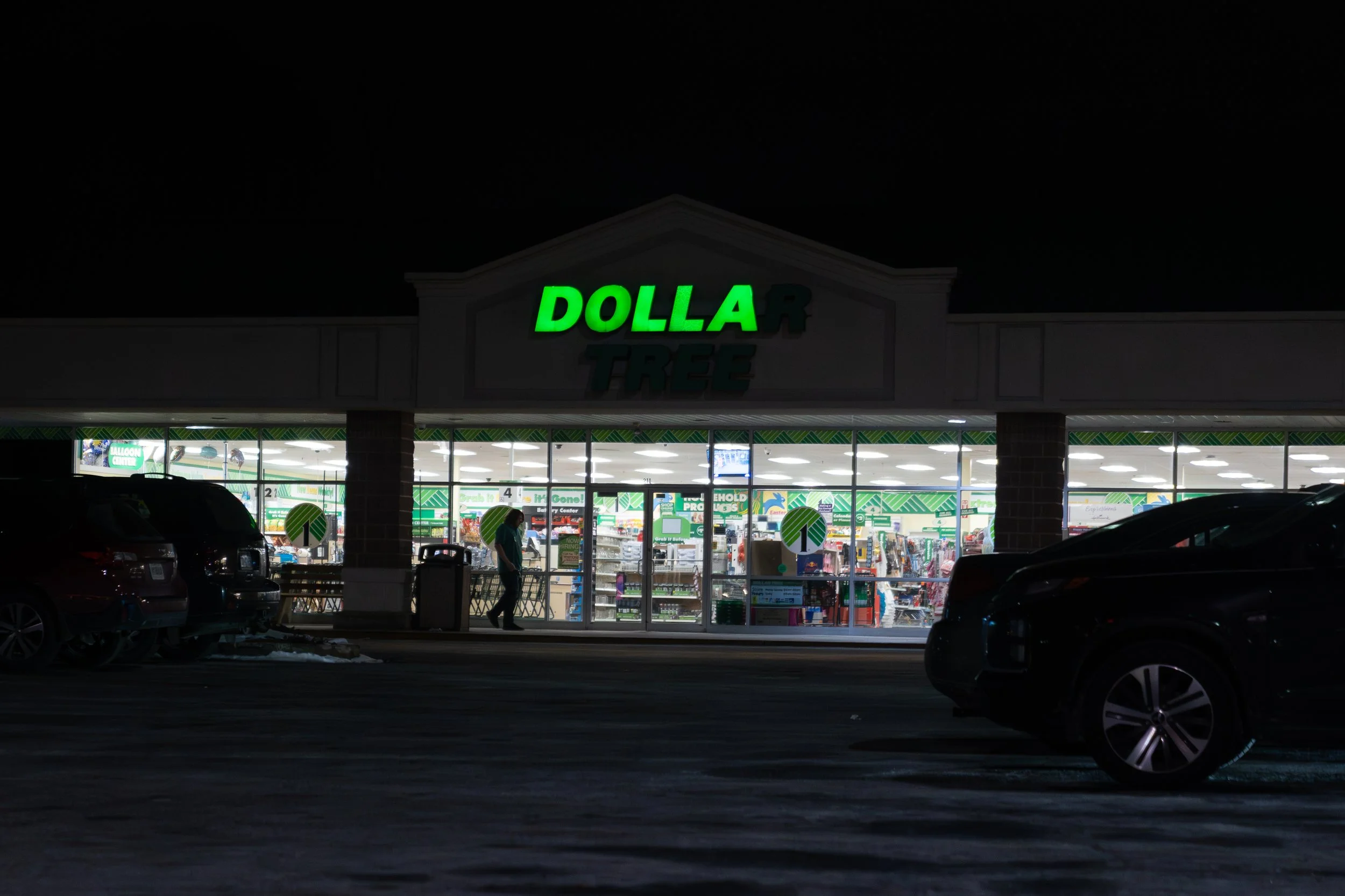 The exterior of a Dollar Tree store at night, with illuminated green sign and visible inside brightly lit shelves and shopping area, several parked cars in the parking lot.