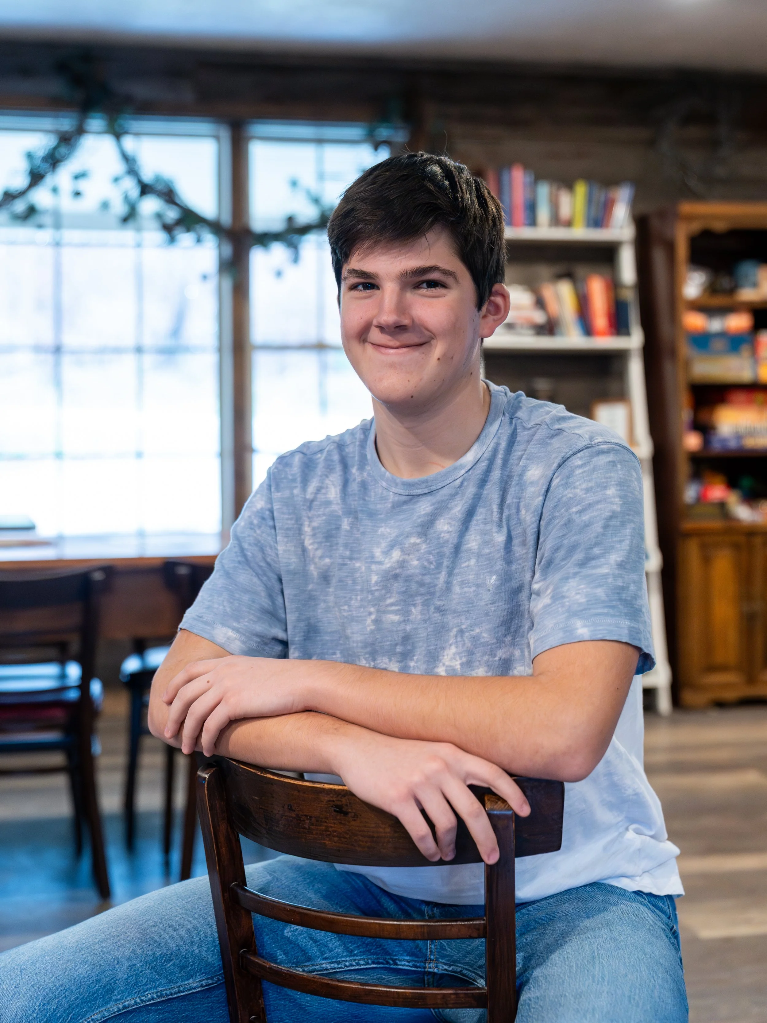 A young man smiles while sitting in a wooden chair with arms crossed in a cozy room with bookshelves and large windows.