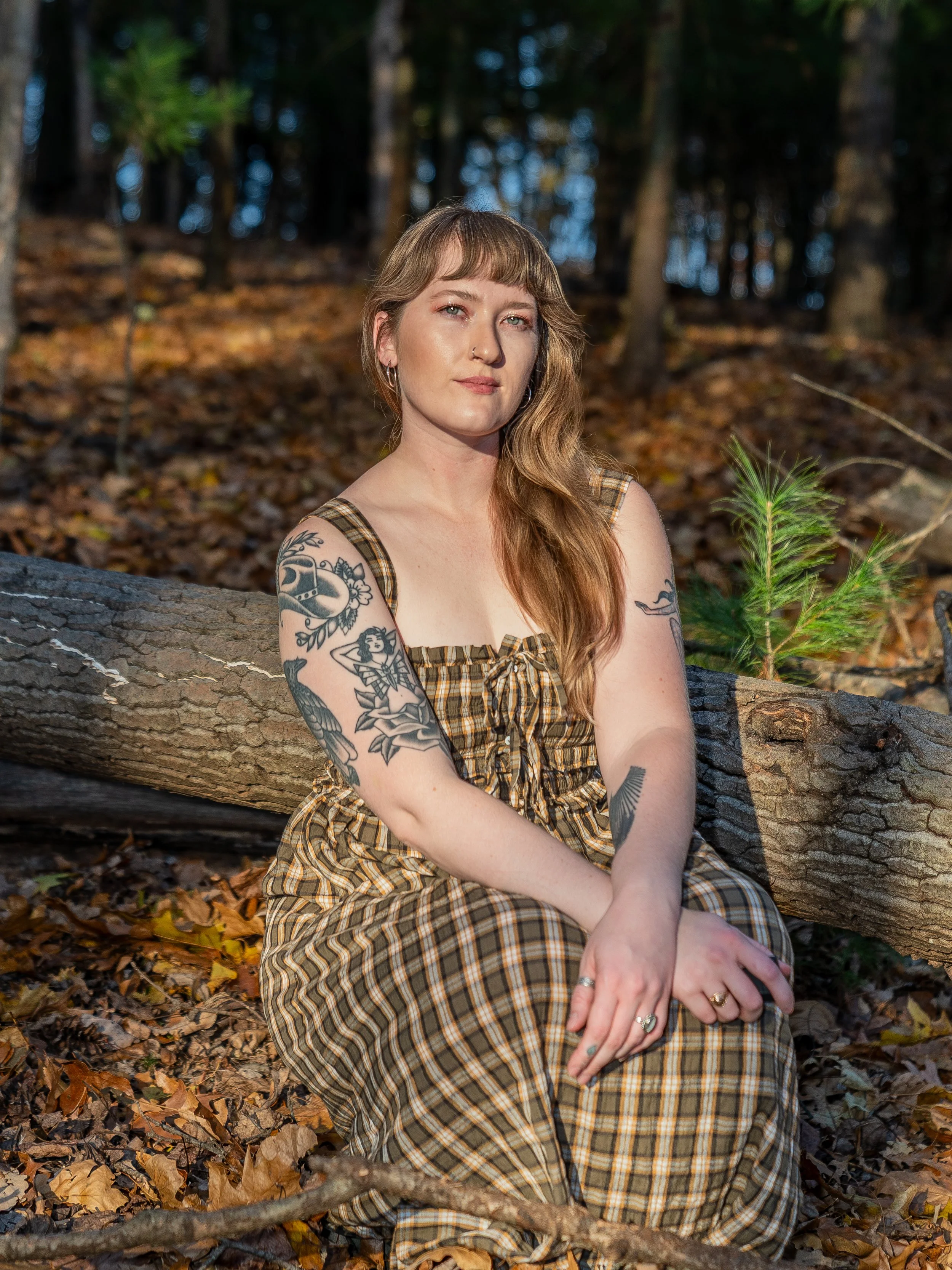 A woman with tattoos on her arms and long wavy hair sitting on a fallen log in a forest, surrounded by autumn leaves, looking at the camera.
