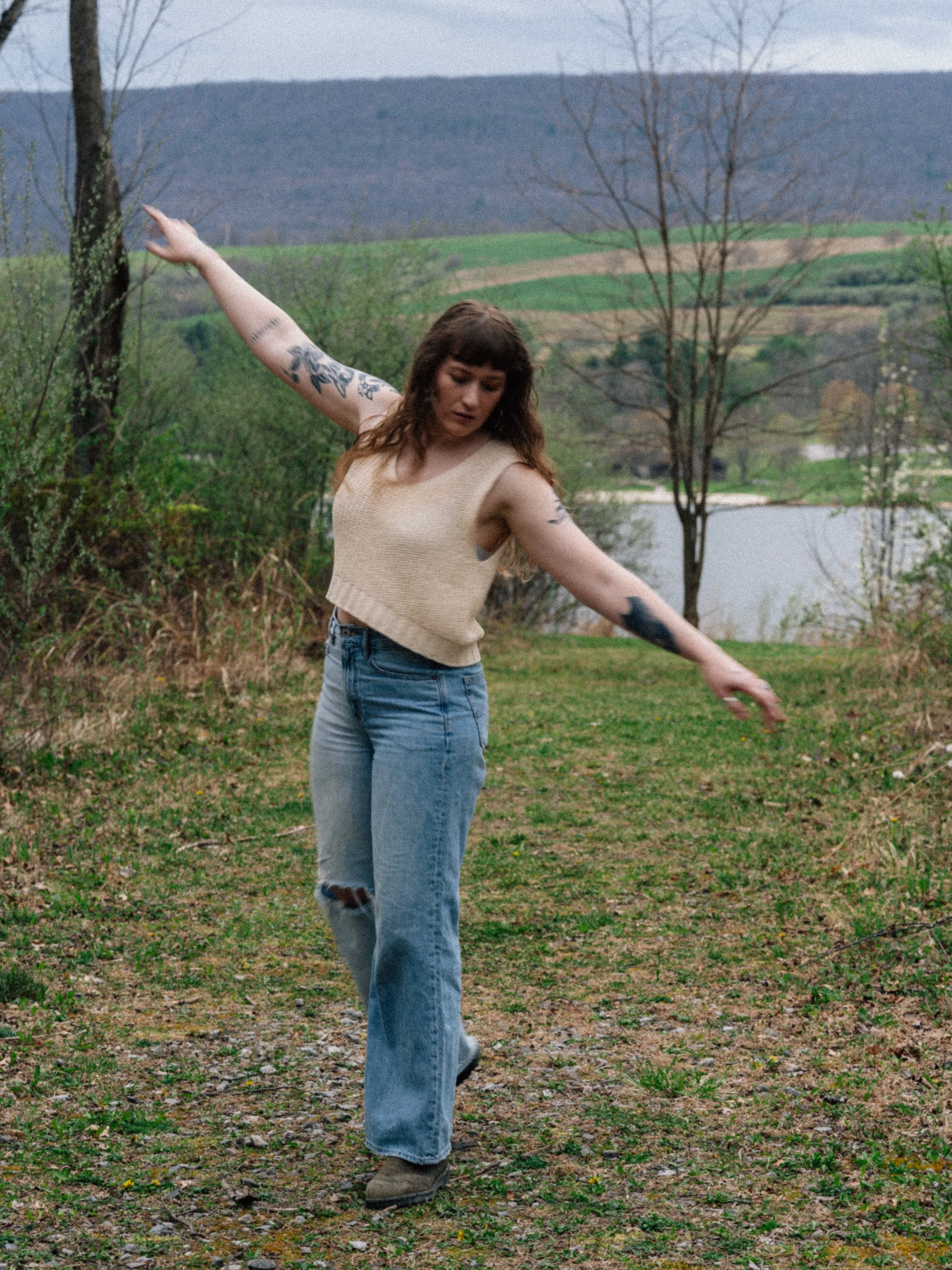 A woman with brown hair and tattoos on her arms, wearing a sleeveless beige knit top and ripped blue jeans, is walking outdoors on a grassy trail with her arms outstretched, in a natural setting with trees, a lake, and distant hills.