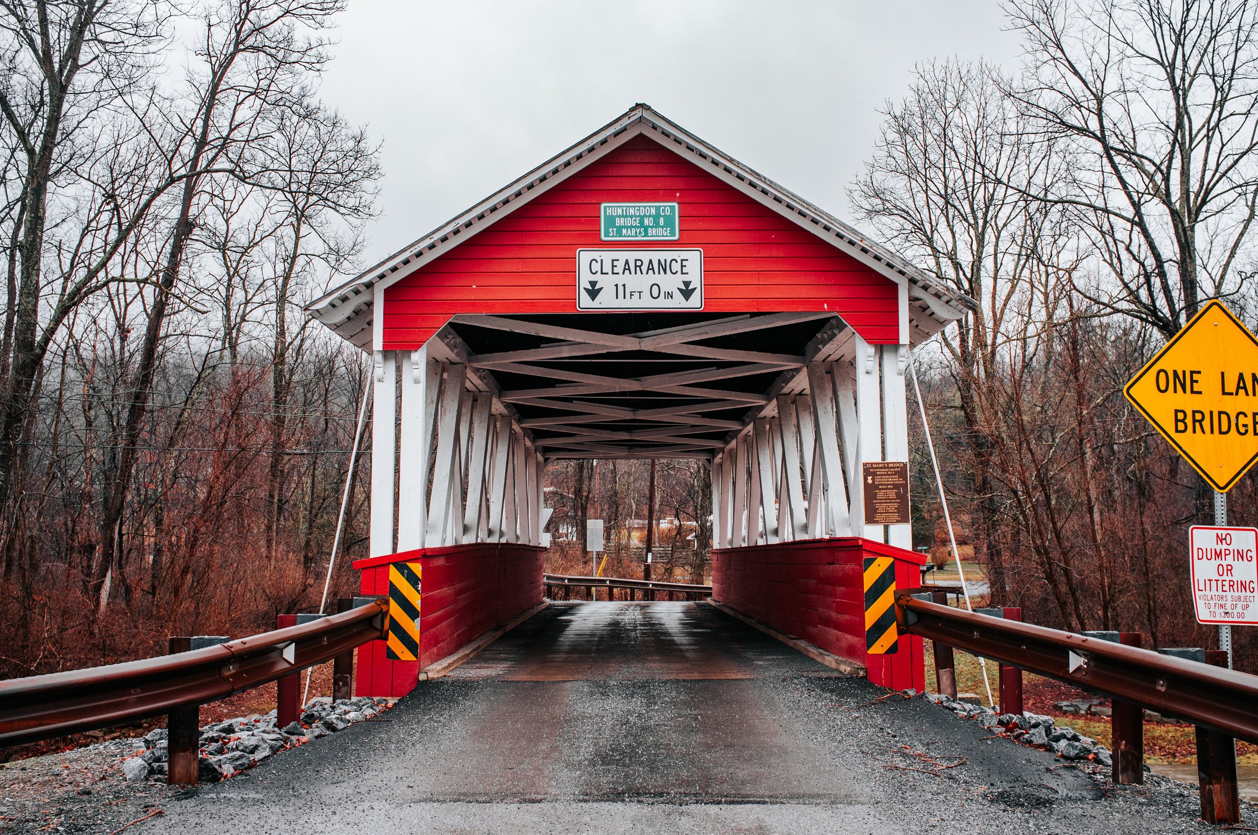 A restored red covered bridge with white framing in a rural setting, with leafless trees and various traffic signs including a height clearance sign and a no dumping sign.